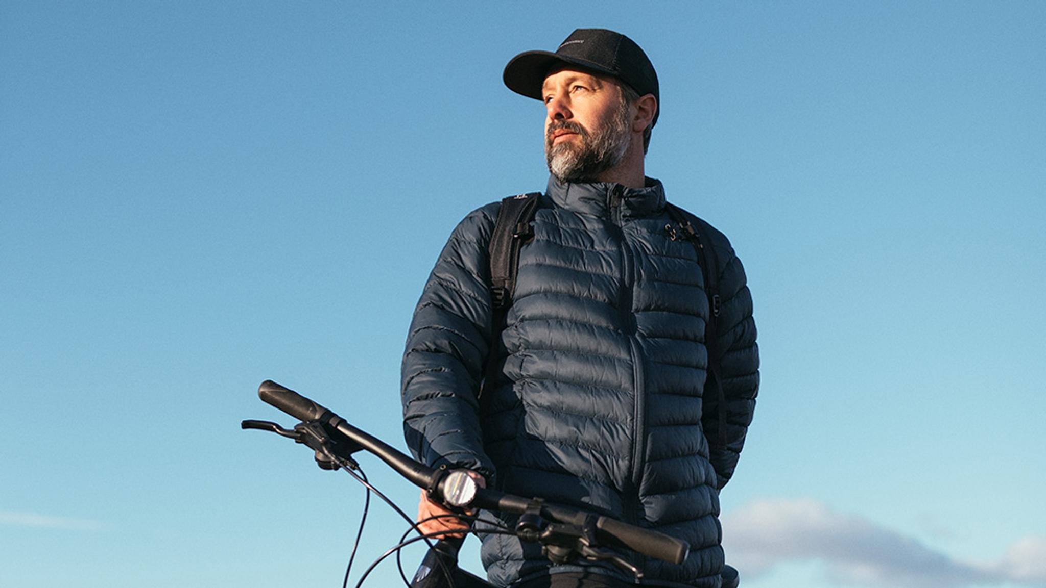 A man in a dark puffy jacket, cap, and backpack holds a bicycle while looking left against a blue sky.