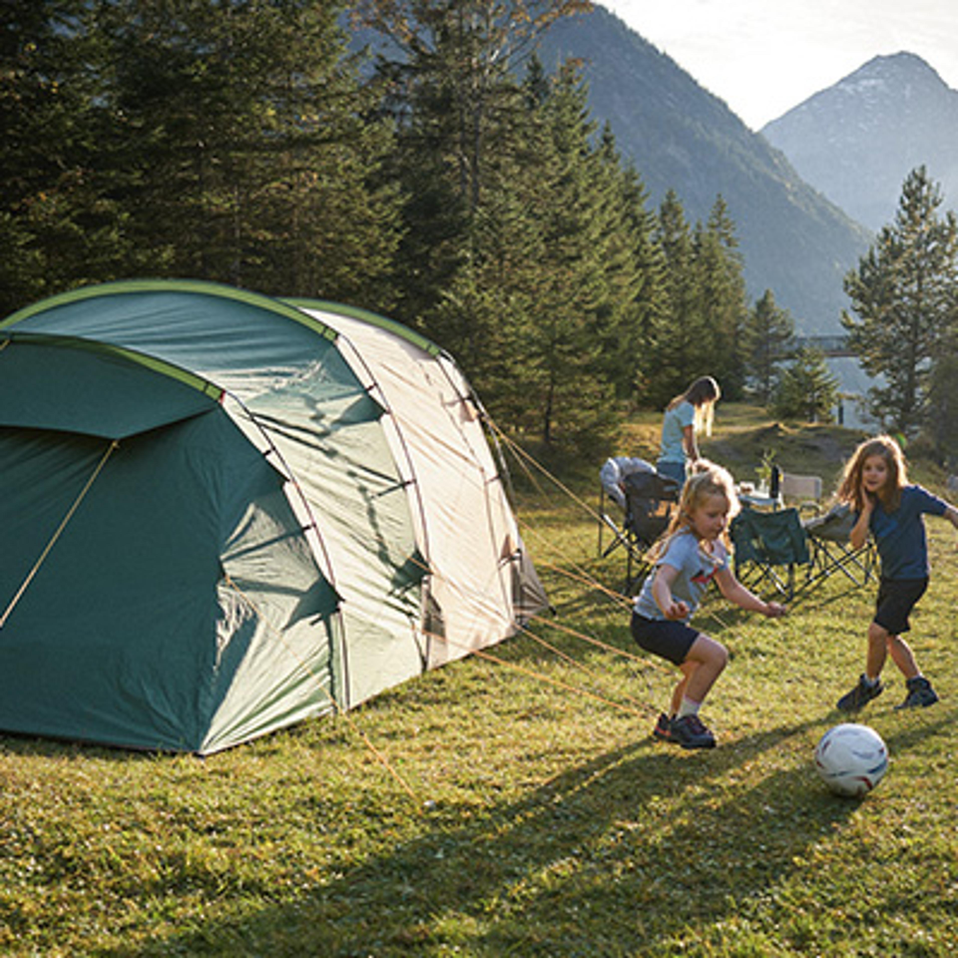 Two children play soccer near a large green tent at a mountain campsite.
