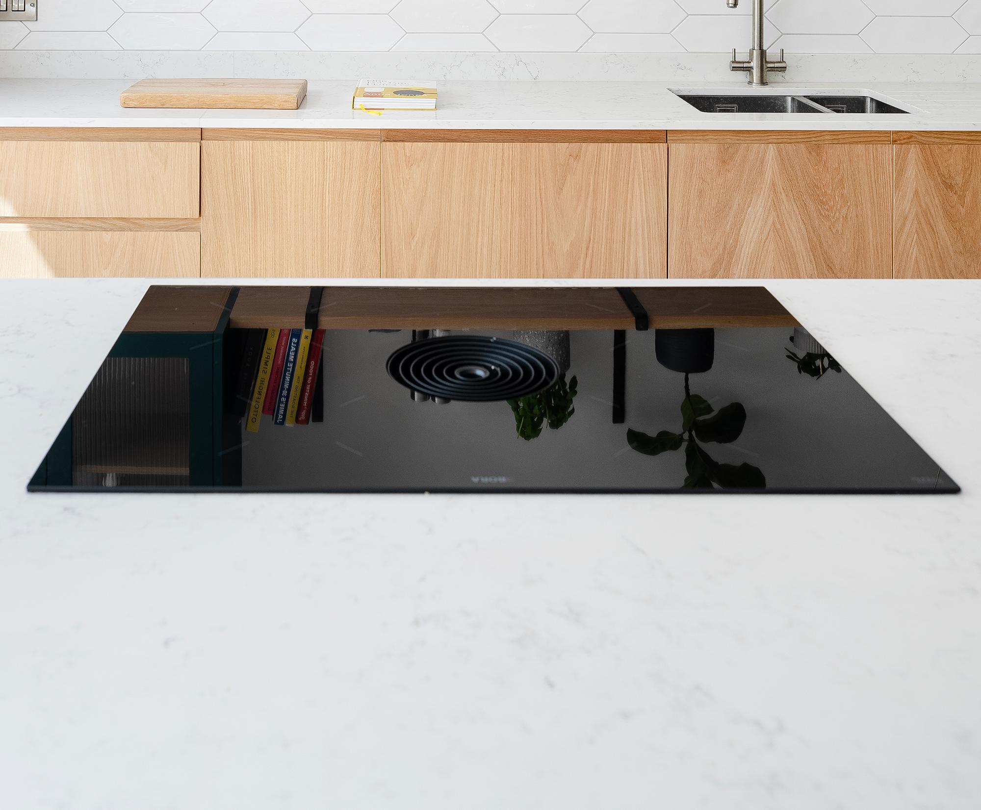 Sleek black induction cooktop with integrated ventilation on a white marble kitchen island, with wooden cabinets, a sink, and hexagonal white tile backsplash in the background.