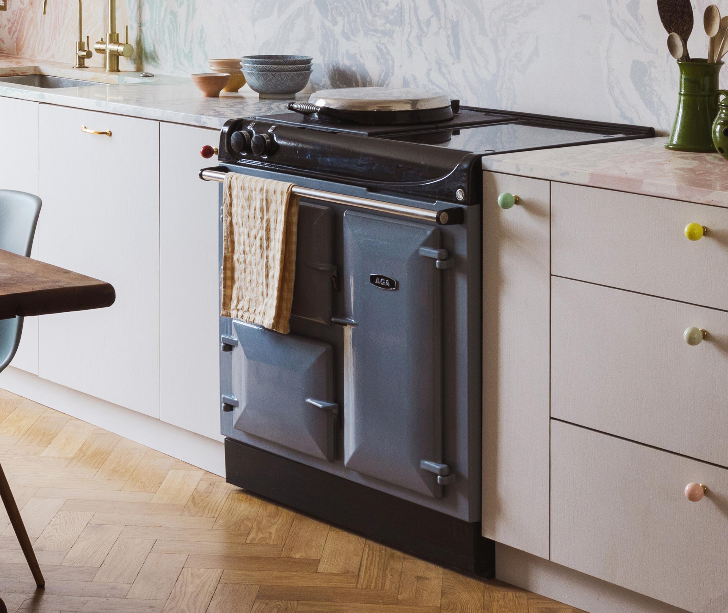 A pastel blue AGA cooker with a beige towel hanging on its handle, set in a modern kitchen with white cabinets, colorful knobs, gold faucet, and a light patterned backsplash.