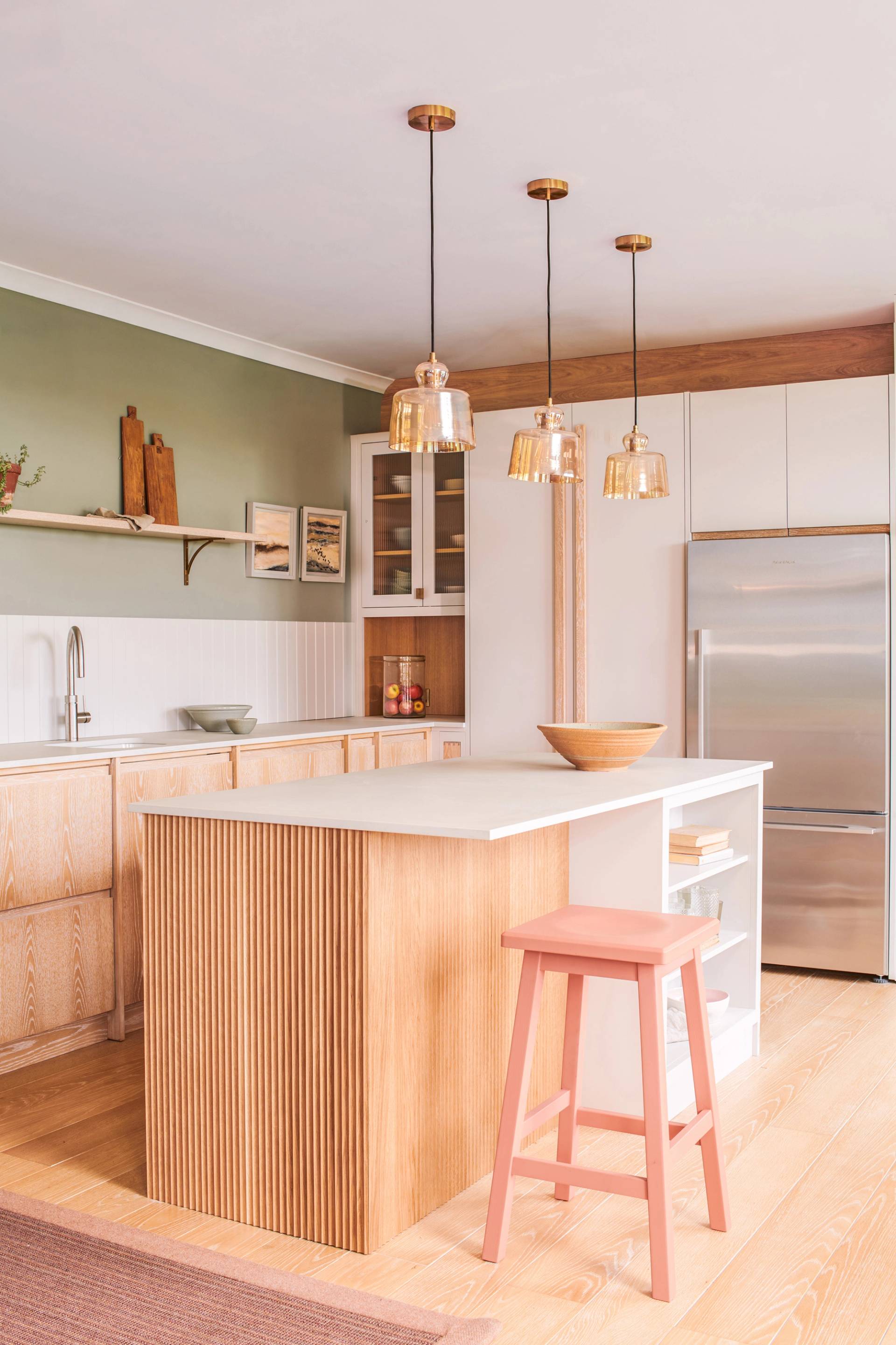 A modern kitchen with light wood cabinets, a large island with vertical wood paneling, a pink stool, pendant lights, white countertops, open shelving, and a stainless steel refrigerator.