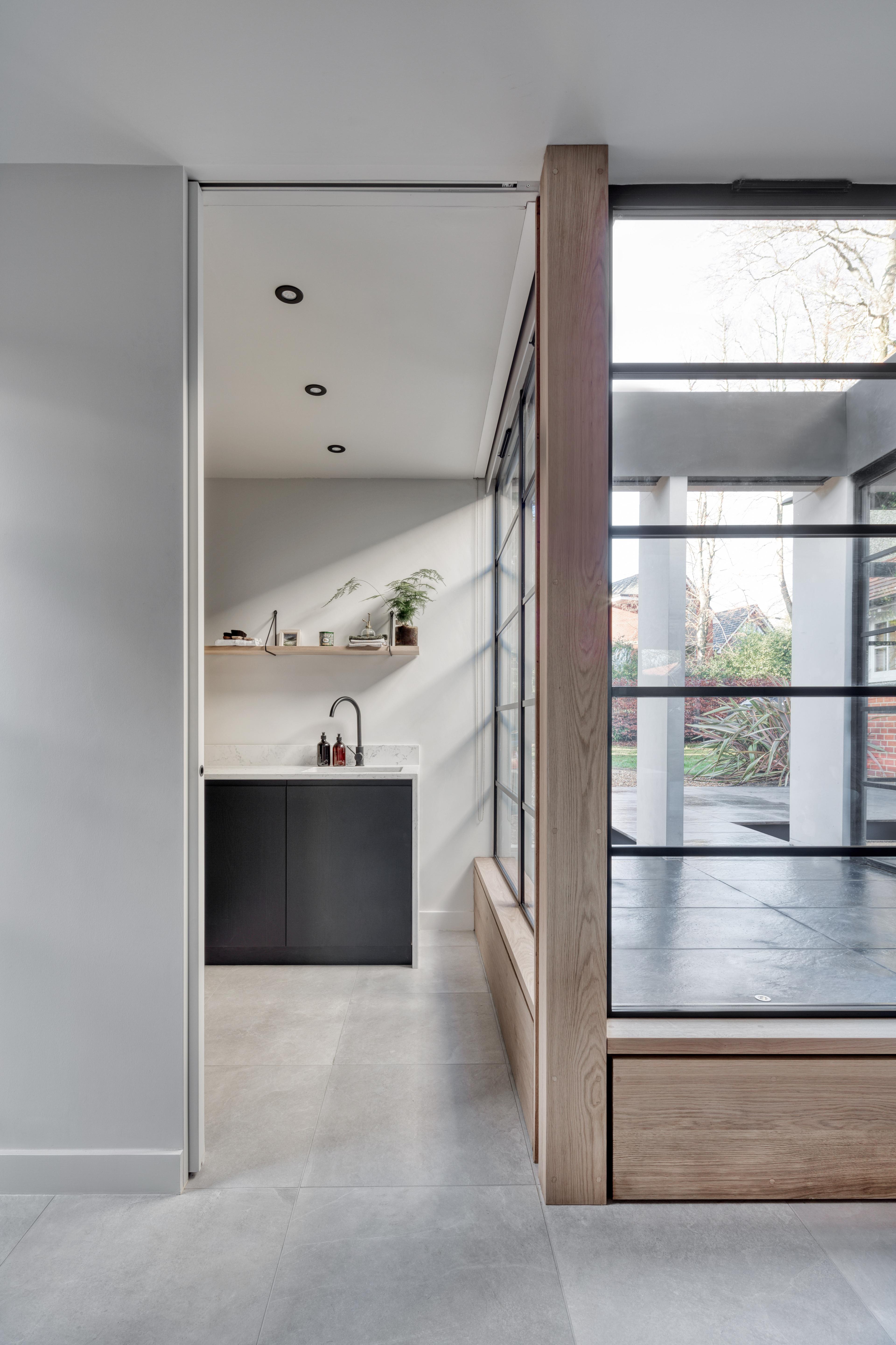 Modern interior featuring a minimalist kitchen with black cabinetry, a white countertop, and potted plants. Adjacent is a sunlit enclosed glass patio area with a view of greenery outside. The space has gray flooring and contemporary design elements