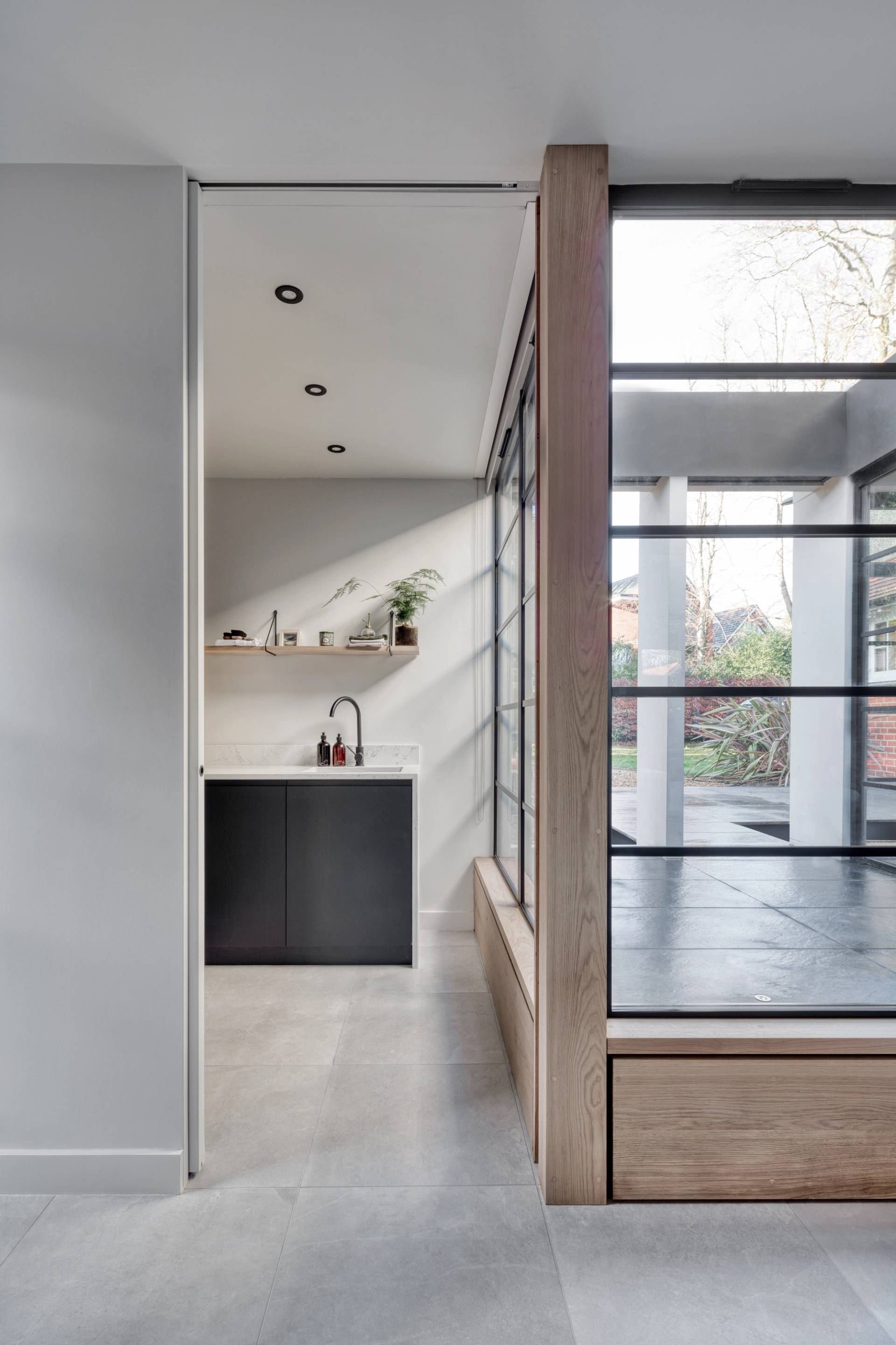 Modern interior featuring a minimalist kitchen with black cabinetry, a white countertop, and potted plants. Adjacent is a sunlit enclosed glass patio area with a view of greenery outside. The space has gray flooring and contemporary design elements