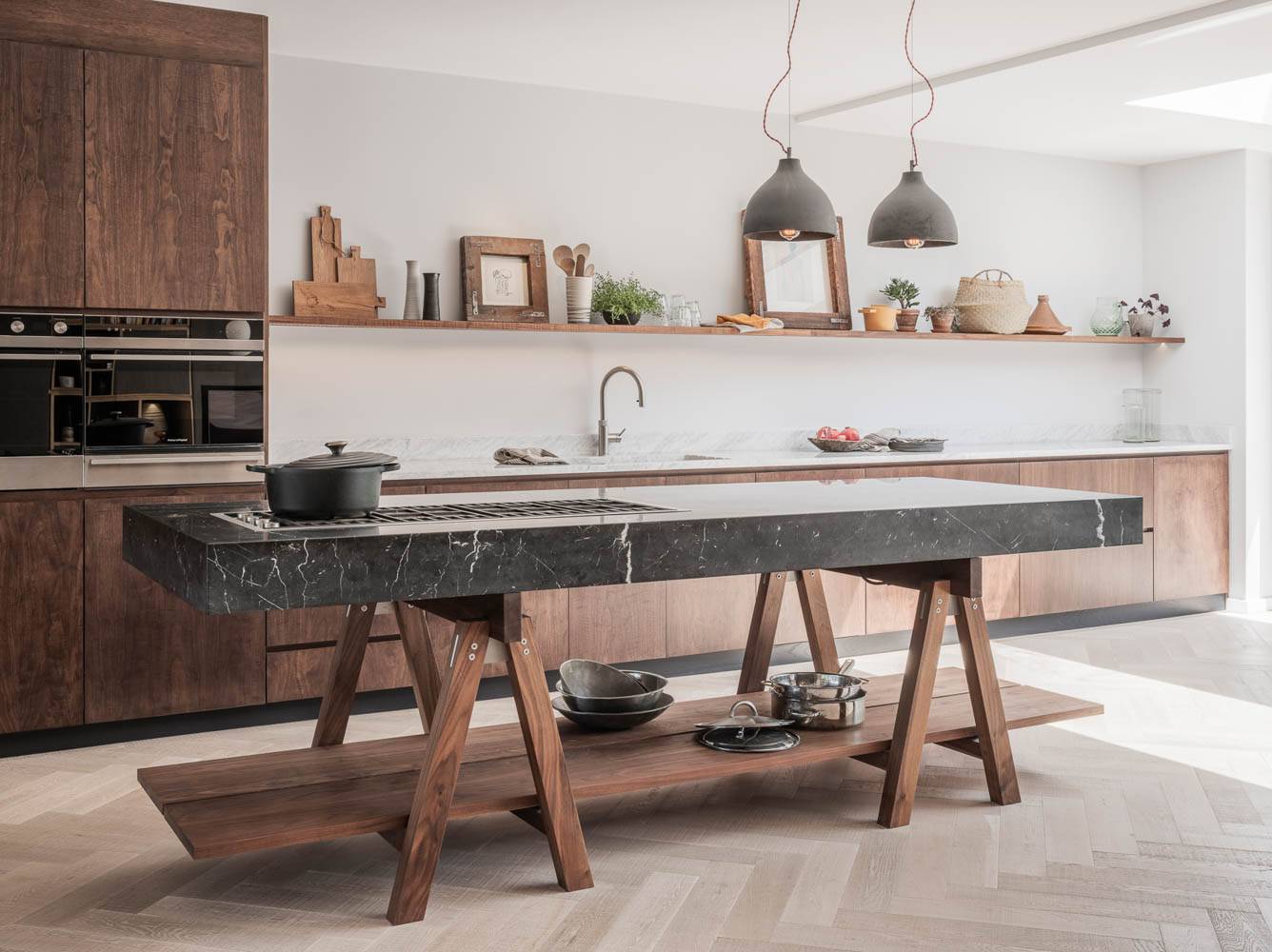 A contemporary kitchen features a dramatic black marble island on a wooden trestle base, set against dark wood cabinetry and a long open shelf with minimalist decor.