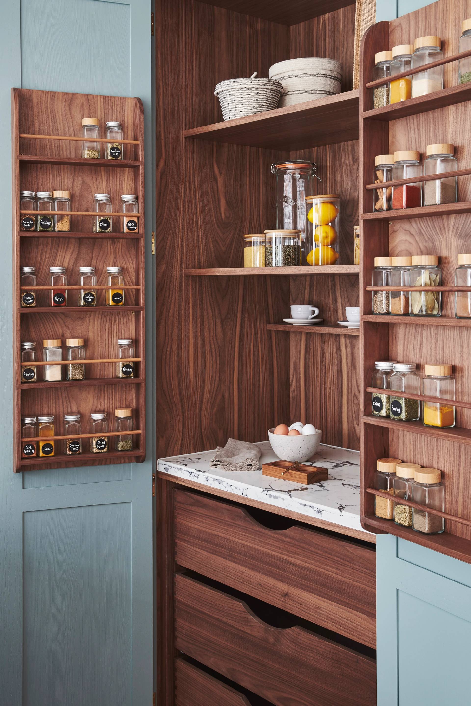 A pantry with light blue doors open to reveal wooden shelves. Spice racks attached to the doors hold various spices in glass jars. Inside are shelves with wicker baskets, a jar of lemons, and cups. A marble counter holds a bowl of eggs.