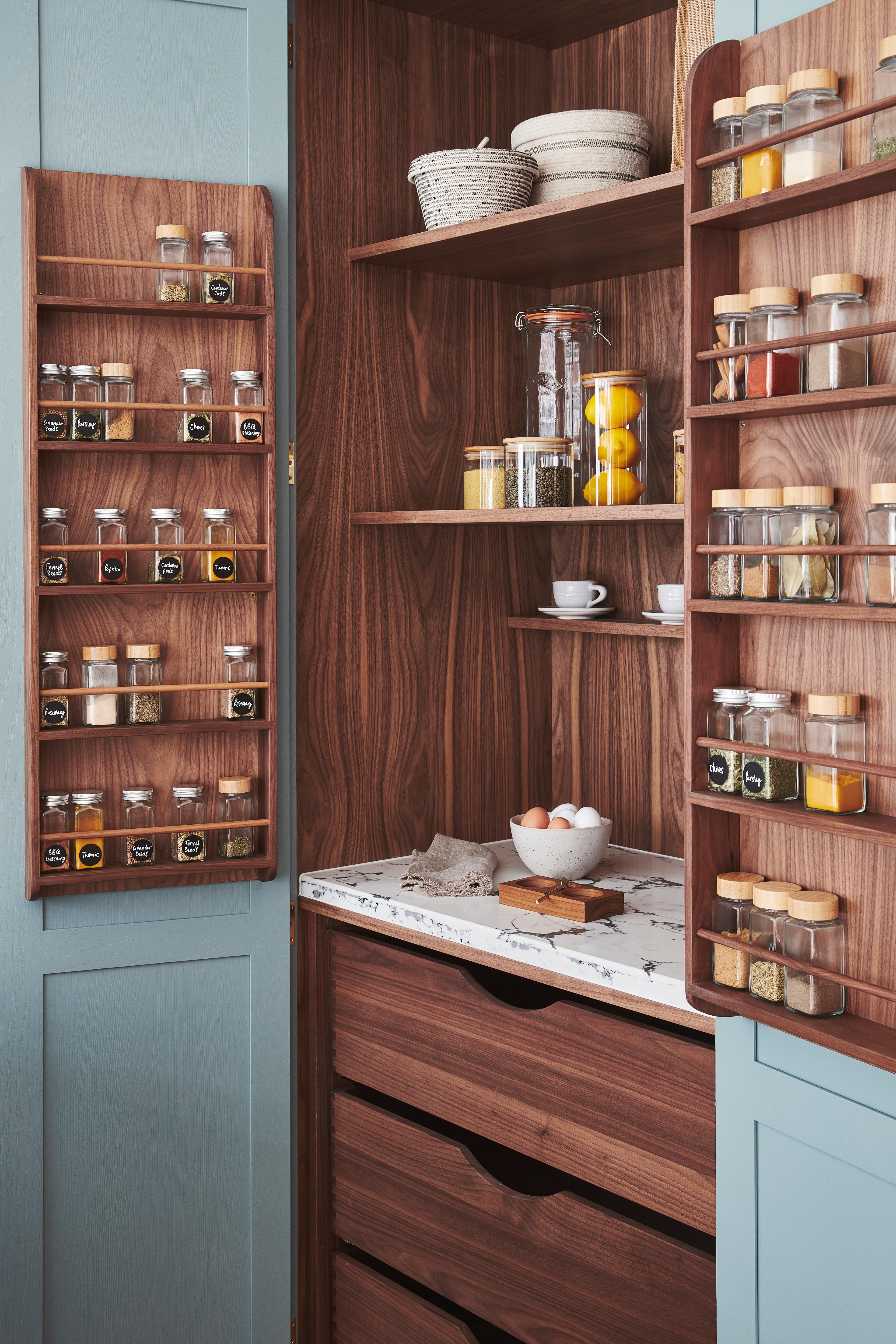 A pantry with light blue doors open to reveal wooden shelves. Spice racks attached to the doors hold various spices in glass jars. Inside are shelves with wicker baskets, a jar of lemons, and cups. A marble counter holds a bowl of eggs.