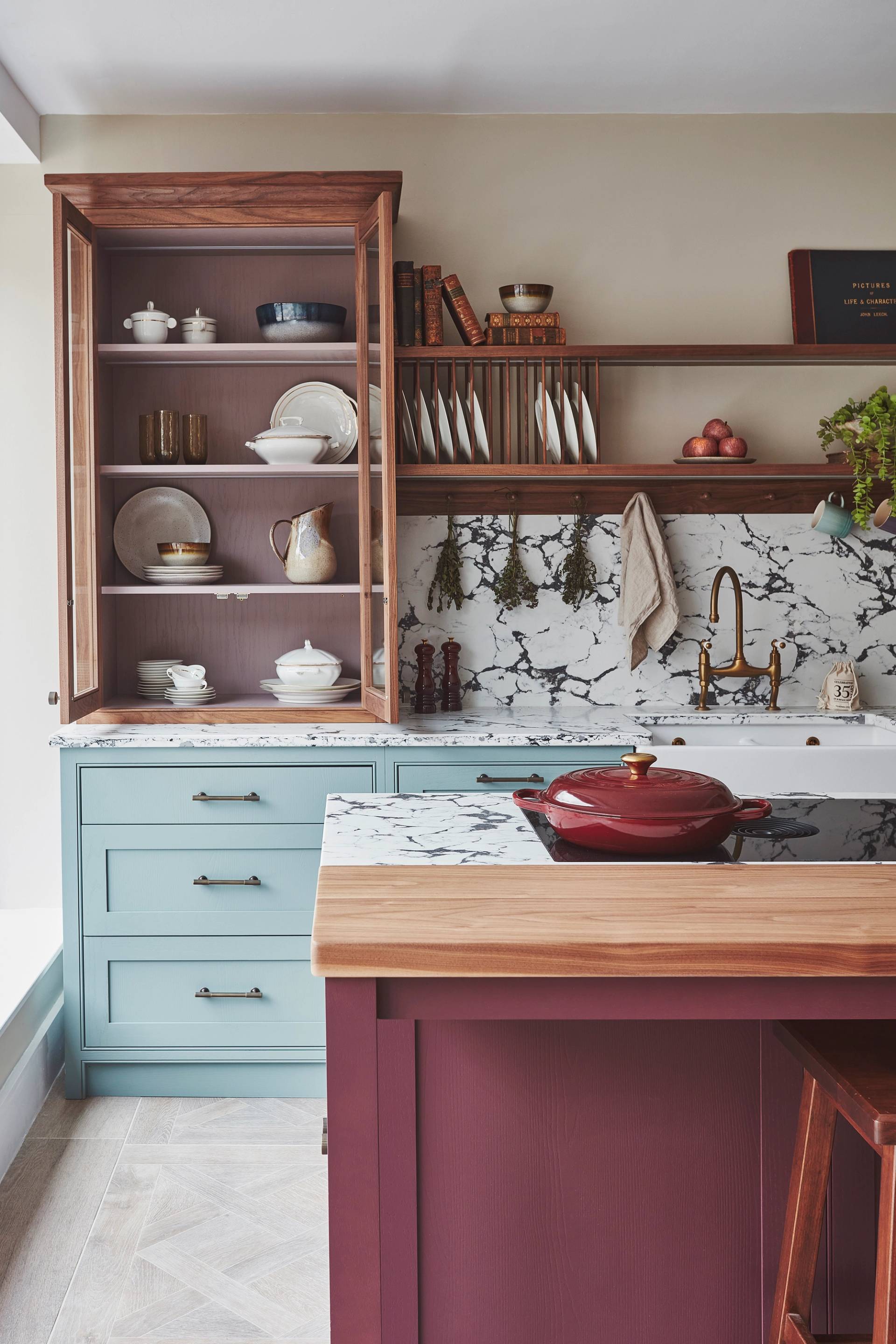 A kitchen with blue cabinets, marble countertops, and a farmhouse sink. Wooden shelves hold dishes, plants, and decorative items. A tall cabinet displays more dishes. A backsplash with a black and white pattern complements the design.