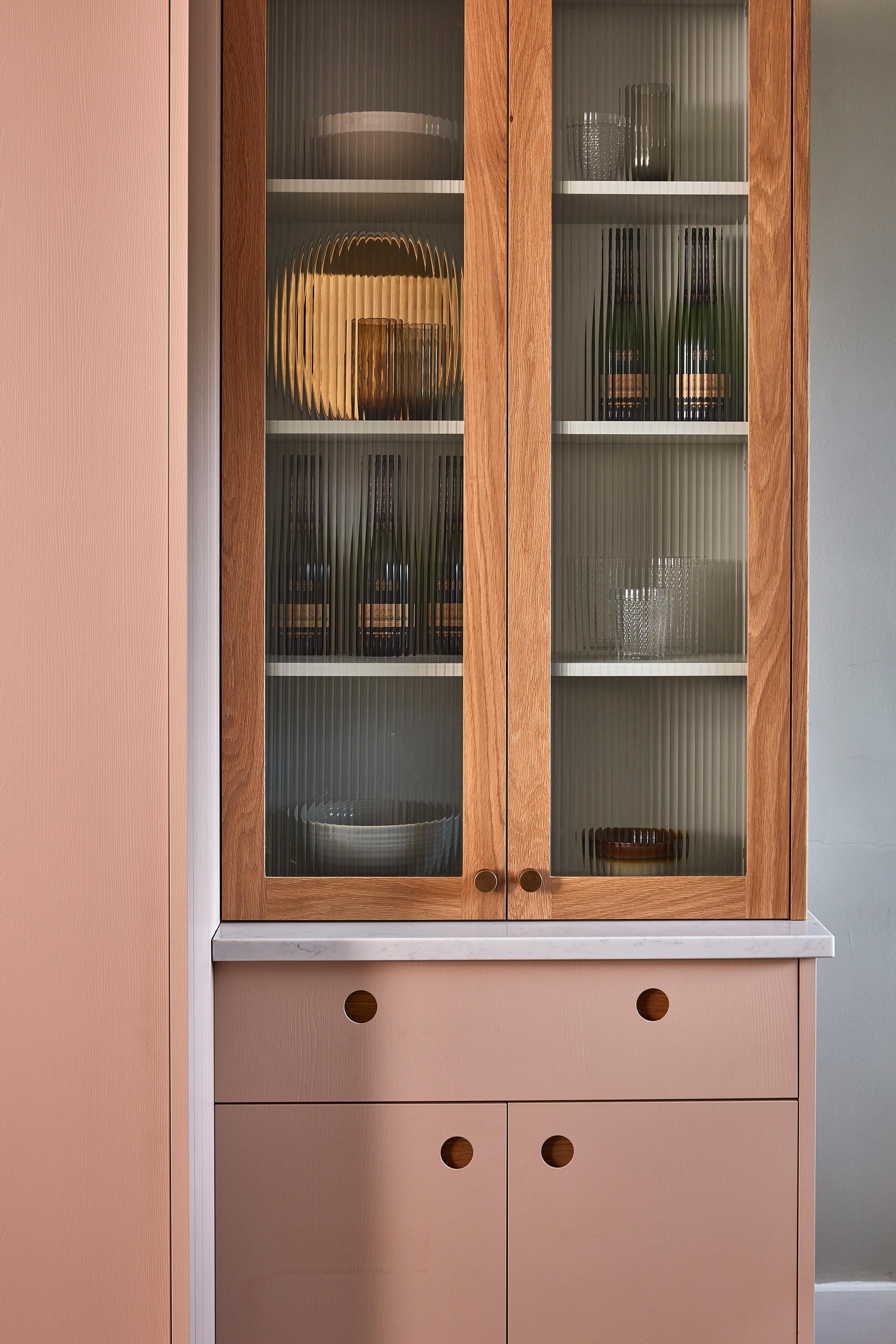 A modern cabinet with ribbed glass doors, wooden frames, and a white countertop. Inside, there are bottles, bowls, and glassware. The blush pink lower drawers have round wooden knobs.