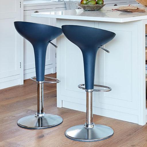 Two modern blue bar stools with chrome bases are placed next to a white kitchen island on a wooden floor. A bowl of green apples sits on the counter in the background.
