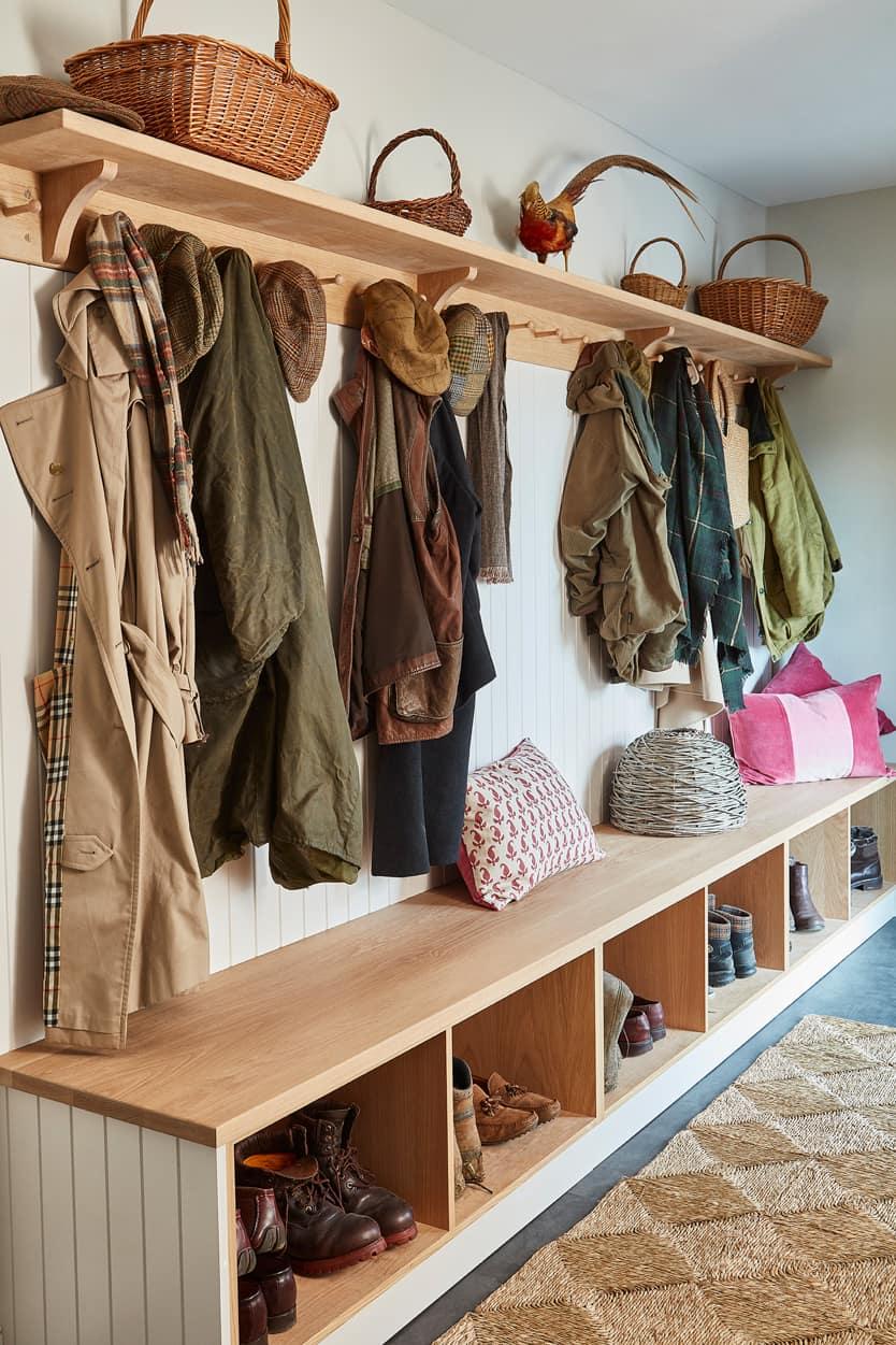 A cozy mudroom with wooden benches and cubbies, coats and hats hanging on wall hooks, baskets on the upper shelf, pillows on the bench, and boots stored below, with a woven rug on the floor.