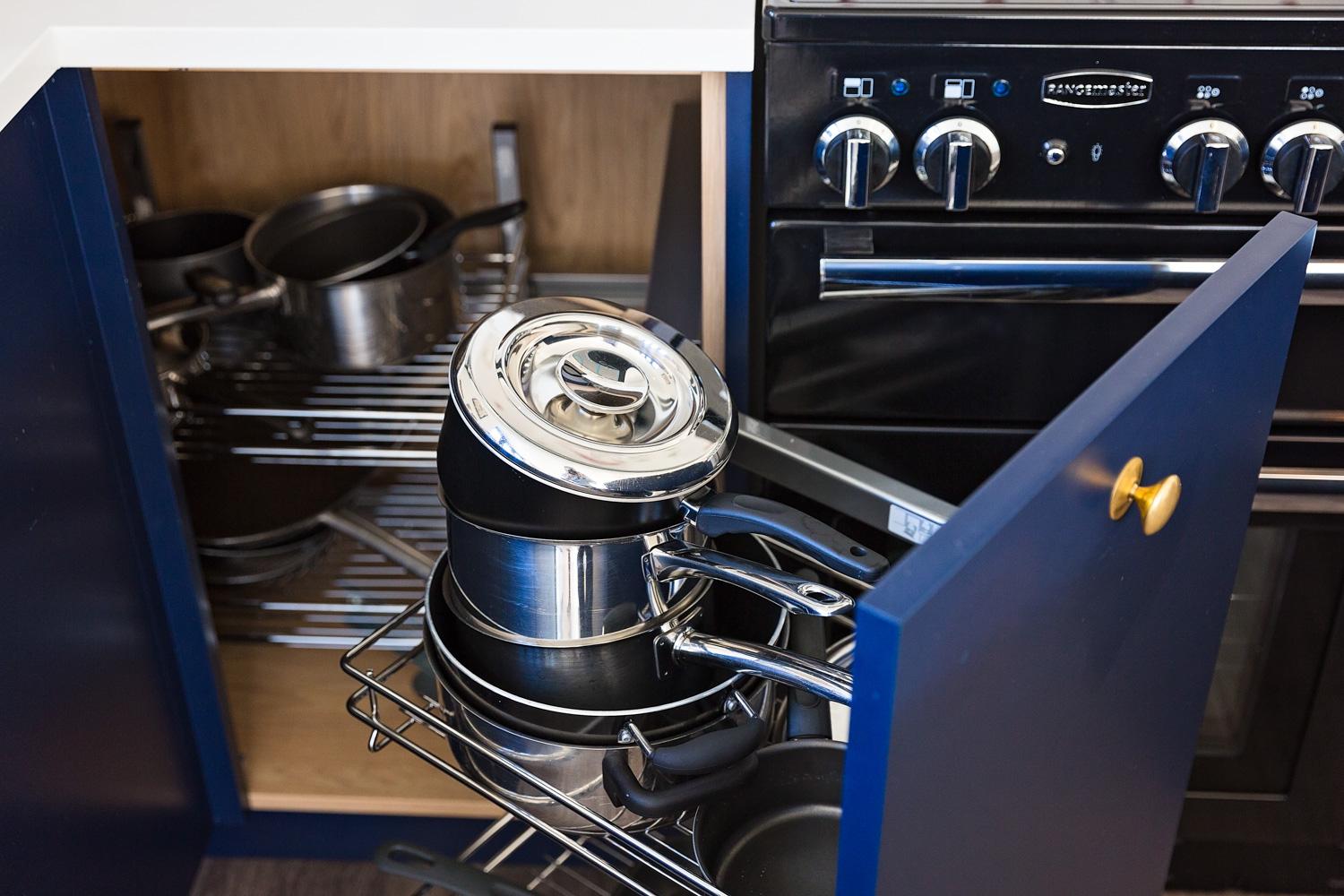 A blue kitchen cabinet with a pull-out wire rack is filled with stacked pots and pans next to an oven with visible control knobs. An adjacent cabinet also stores cookware.