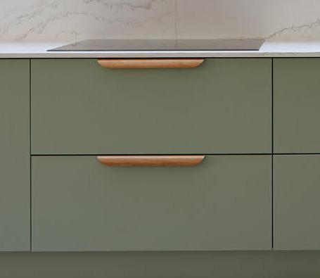A close-up of sage green kitchen drawers with light wooden handles, set against a white countertop and a light marble-patterned backsplash.