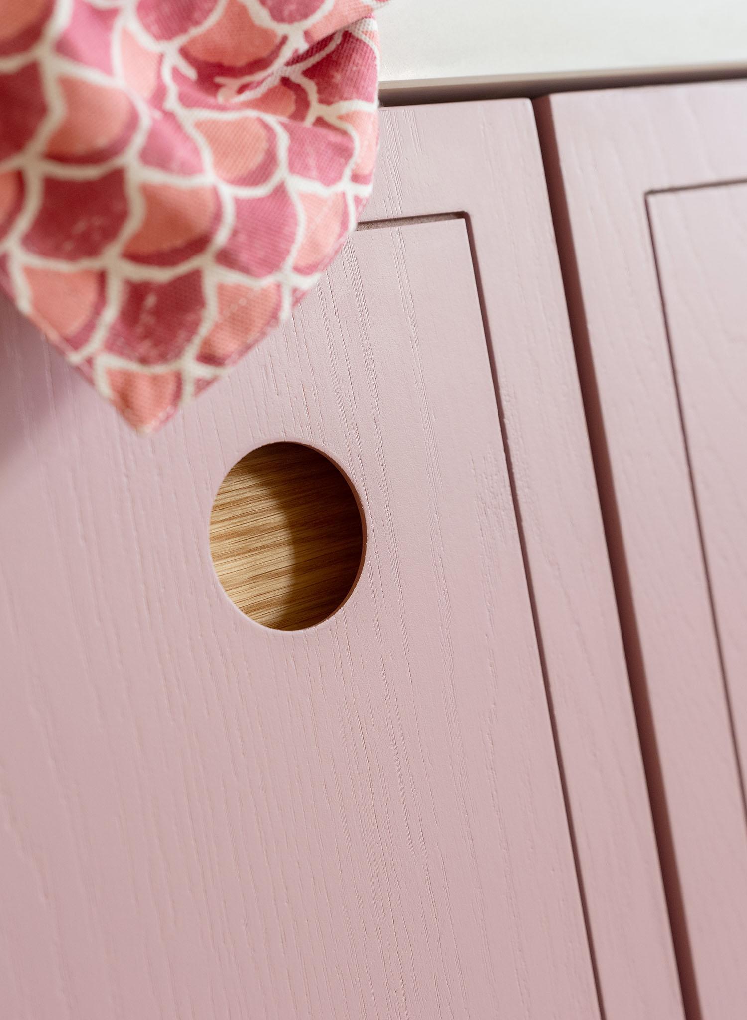 A close-up of a pink wooden cabinet with a round finger hole handle, revealing a glimpse of light wood inside. A pink and white patterned cloth is draped over the top edge.