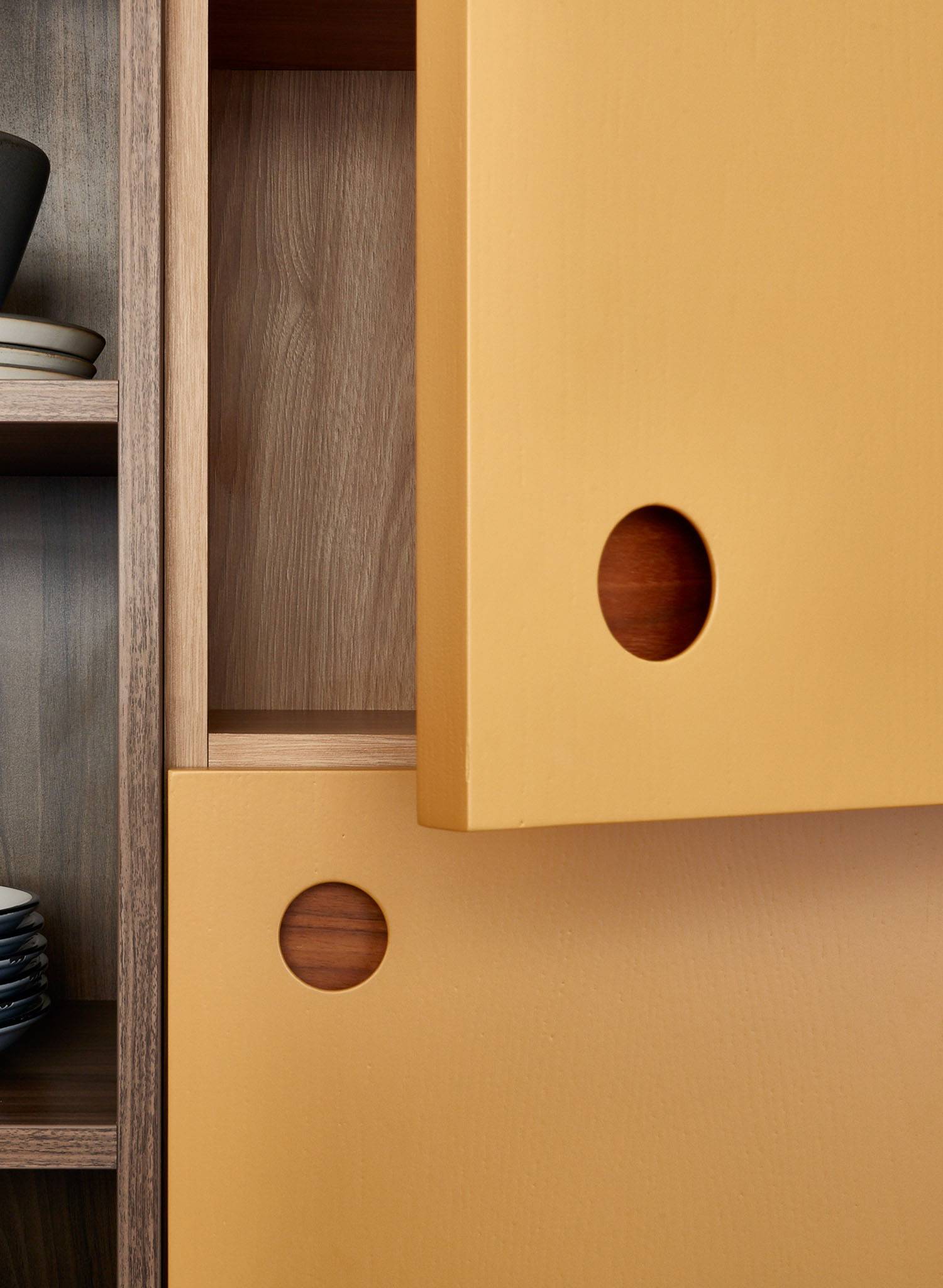 Close-up of a wooden cabinet with yellow sliding doors featuring circular cut-out handles, partially open to reveal the wood grain interior. Shelves on the left hold stacked plates and a bowl.