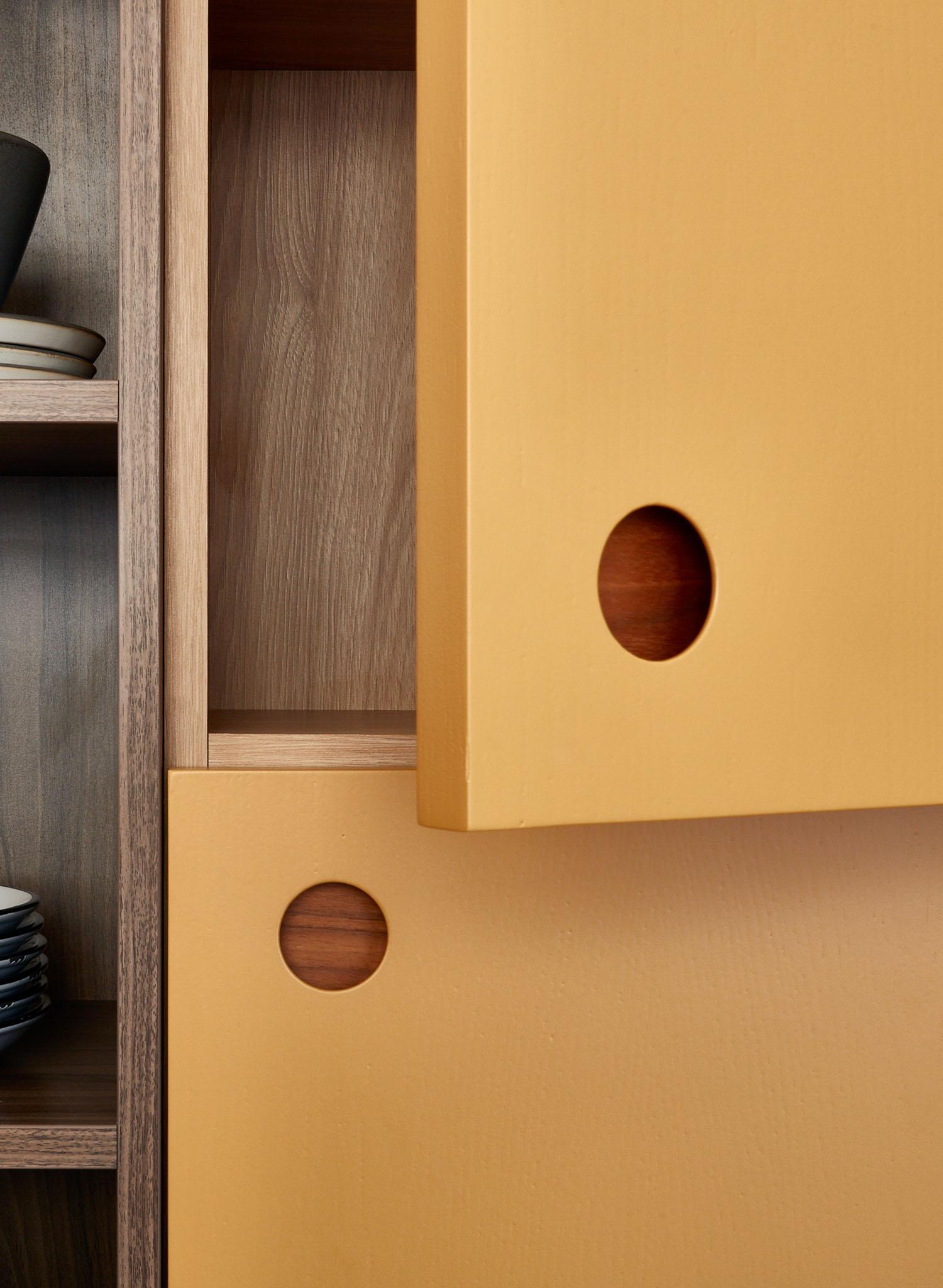 Close-up of a wooden cabinet with yellow sliding doors featuring circular cut-out handles, partially open to reveal the wood grain interior. Shelves on the left hold stacked plates and a bowl.