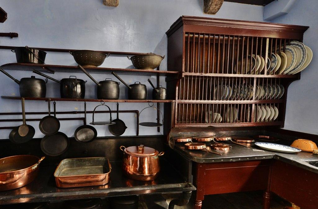 A historic kitchen interior showcasing a large wooden plate rack filled with ceramic dishes next to wall-mounted shelves holding black cast iron pots, pans, and strainers, with an assortment of polished copper cookware arranged on the countertops below.