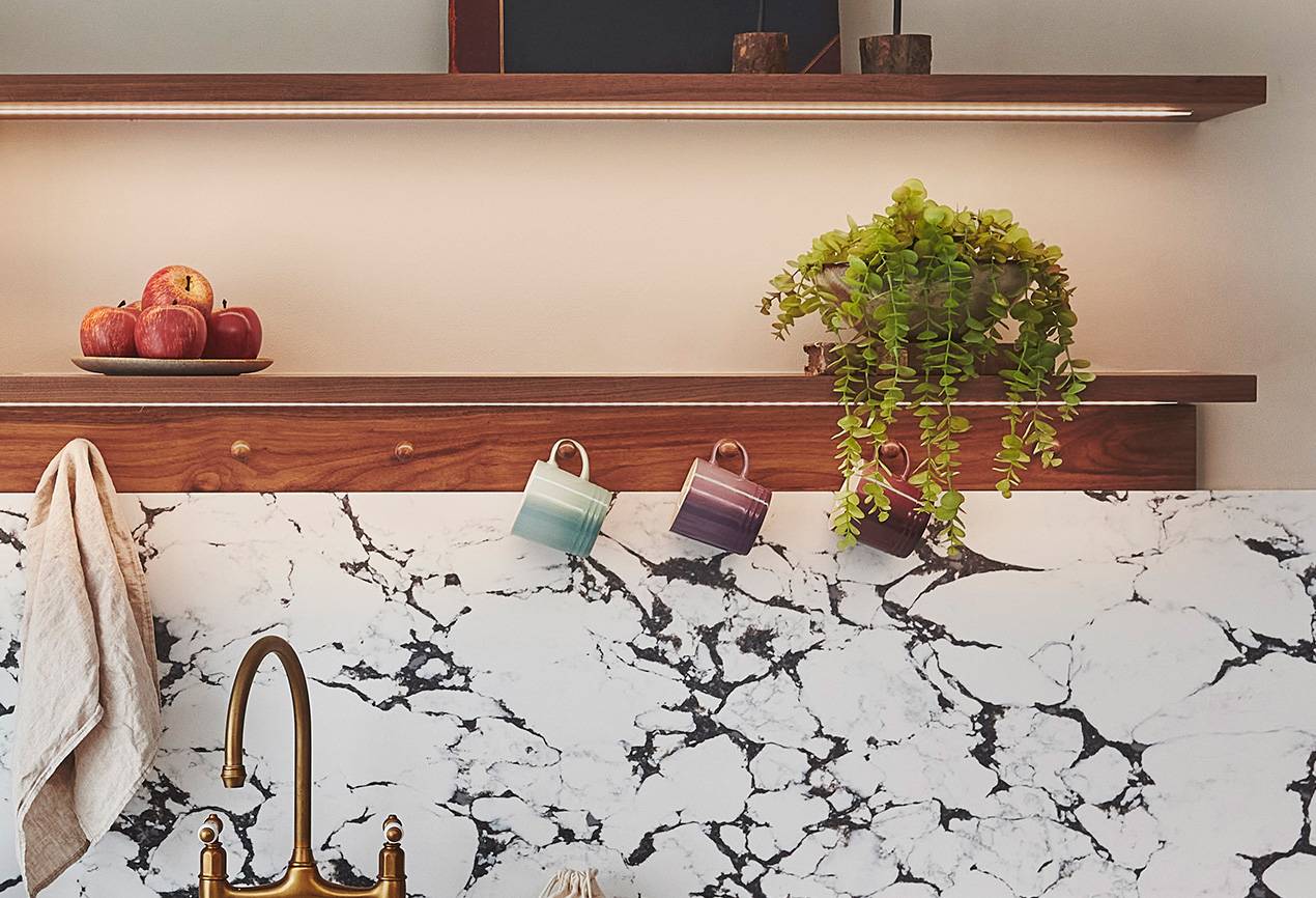 Kitchen scene with a marble-patterned backsplash, a golden faucet, and hanging towels. A wooden shelf holds two pastel mugs and a plant. A top shelf displays a bowl of apples and small decor items against a light-colored wall.