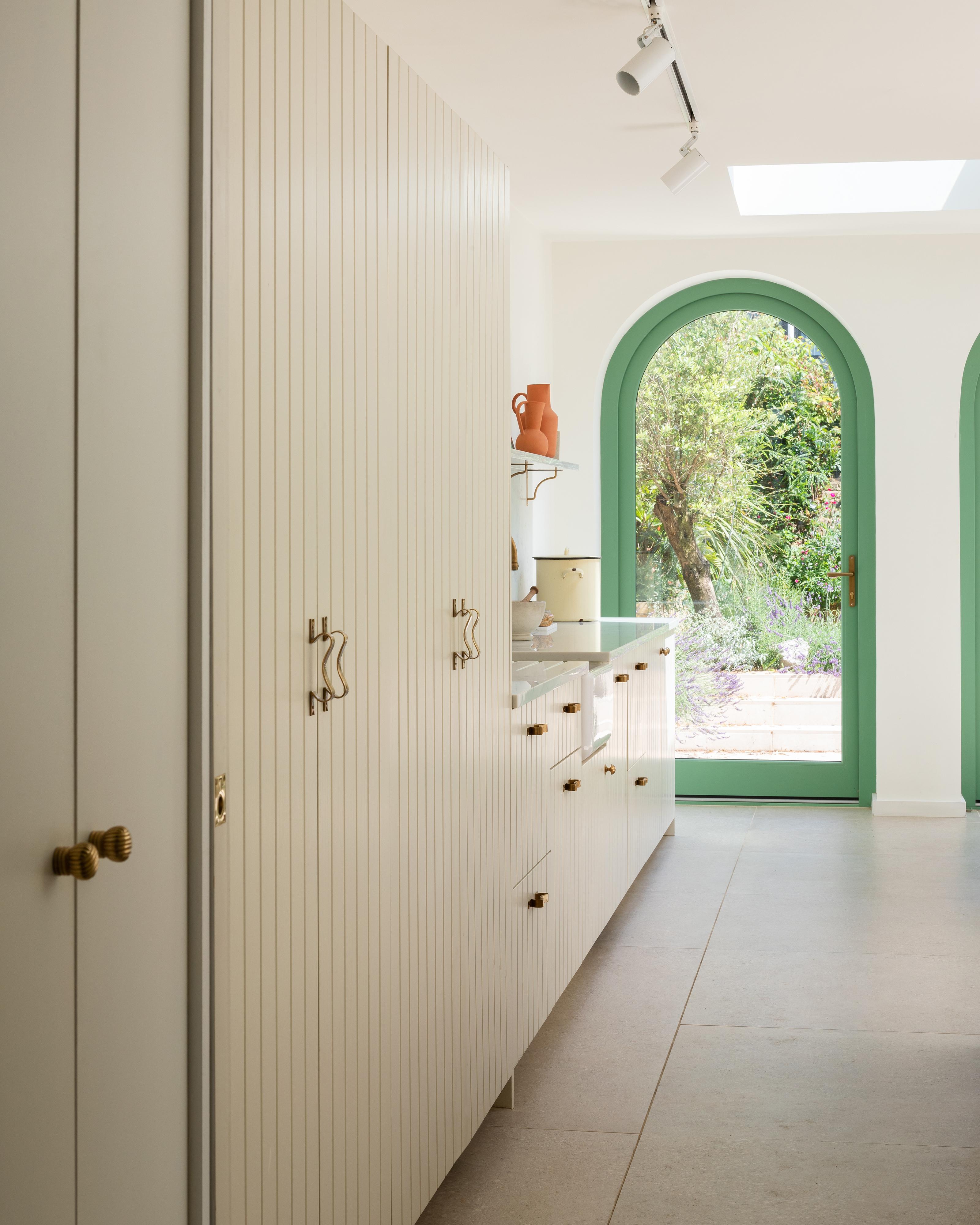 A modern kitchen with cream-colored cabinets, brass handles, and a light beige floor. Large arched glass doors with green frames open to a lush garden outside, and natural light fills the space.