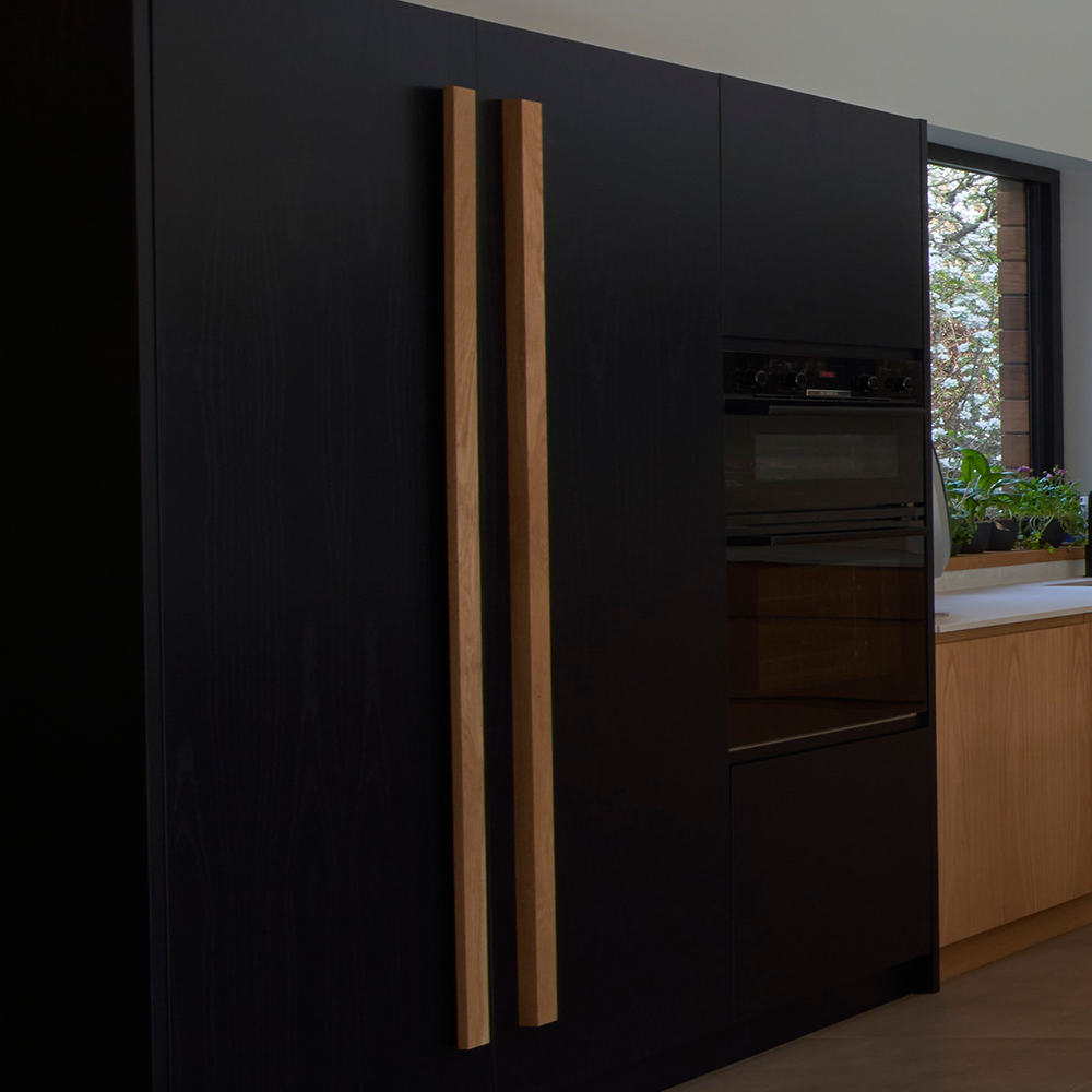 A modern kitchen featuring sleek black cabinets with tall, light wood handles, a built-in oven, and a window above the sink with potted plants on the windowsill.