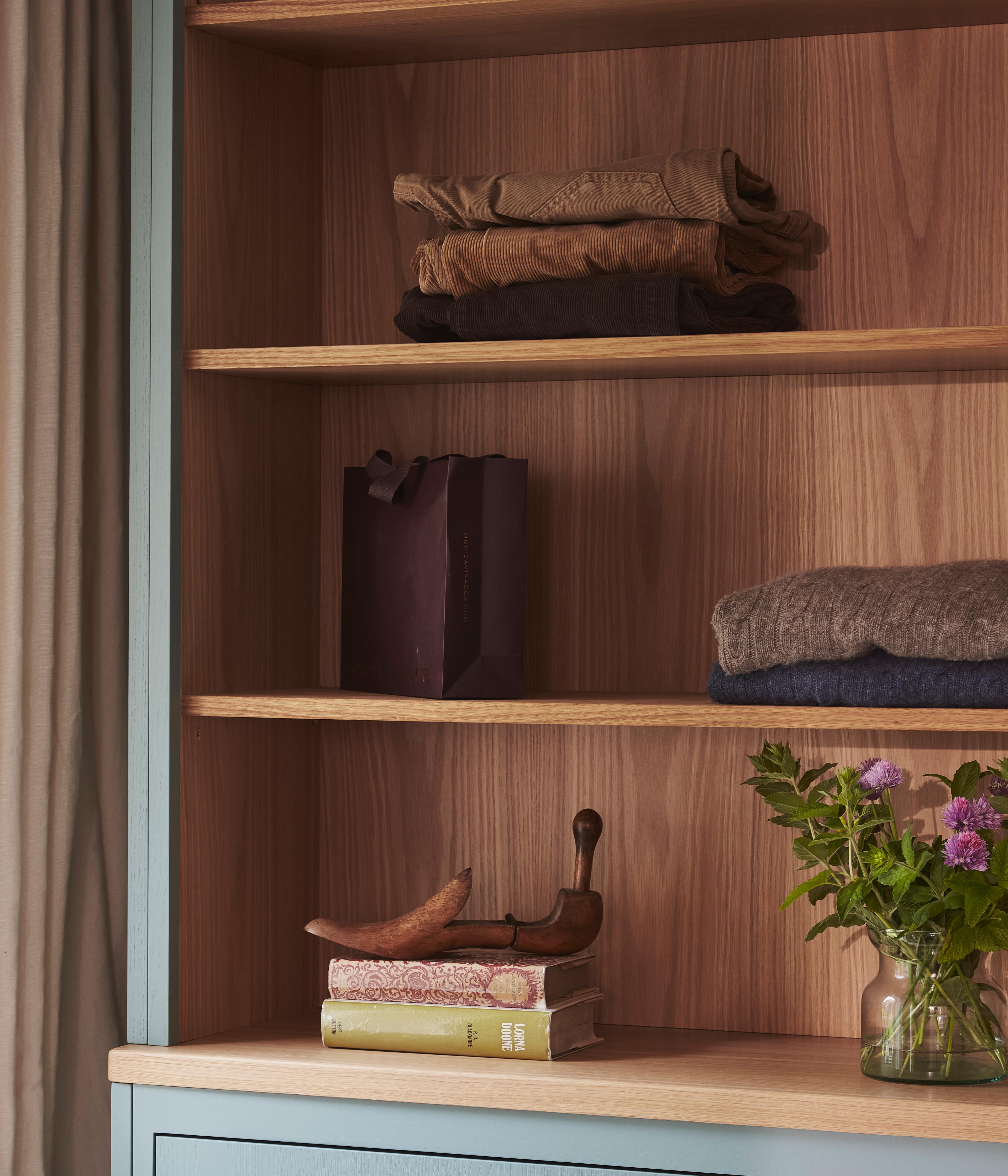 A wooden bookshelf with folded clothes and a paper bag on the shelves. On the lower shelf are stacked books, a wooden decorative object, and a vase of purple flowers. The shelf has a blue frame and wood interior.