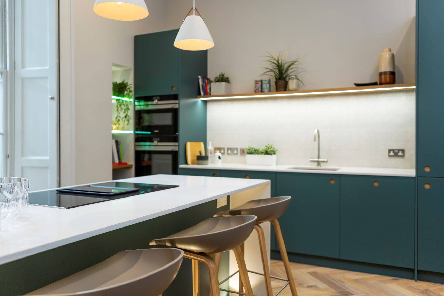 A modern kitchen with teal cabinets, a white countertop island, and three stools. Pendant lights hang above the island, and a plant and small decor items are placed on the upper shelf. The room is well-lit with natural and ambient lighting.