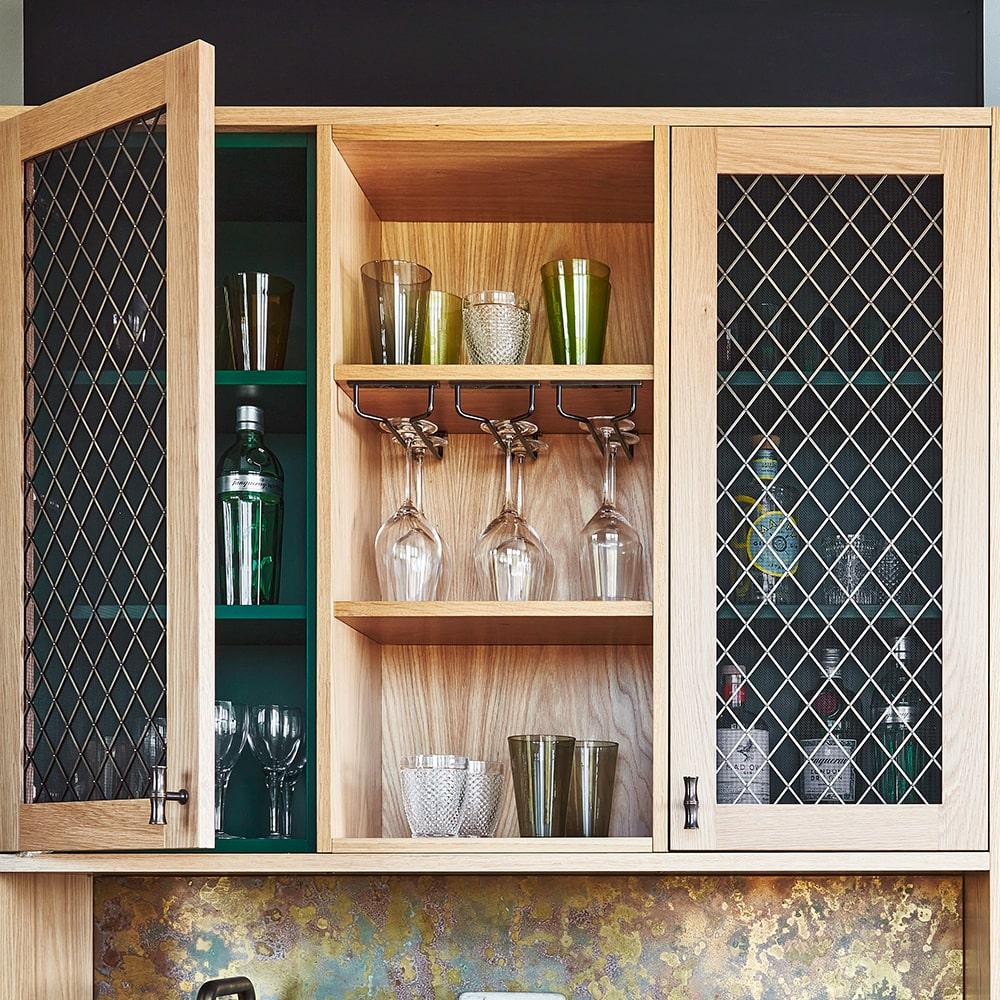 Open wooden cabinet with wire mesh doors displaying glassware, including wine glasses, tumblers, and bottles, organized on shelves; some glasses hang upside down from a rack inside the cabinet.