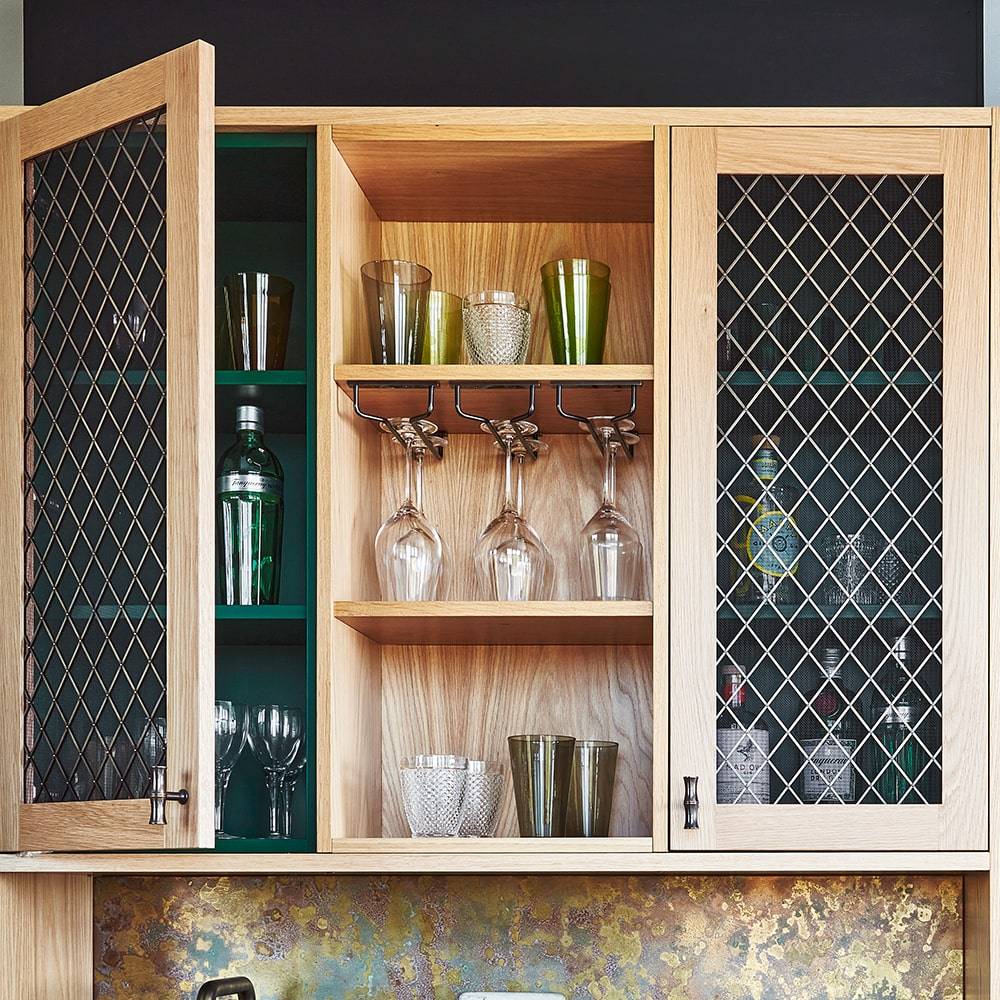 Open wooden cabinet with wire mesh doors displaying glassware, including wine glasses, tumblers, and bottles, organized on shelves; some glasses hang upside down from a rack inside the cabinet.