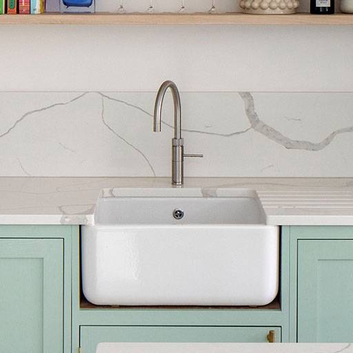 Light blue kitchen cabinets with gold handles, a white countertop, a tall green plant, and wicker bar stools at a marble-topped island in a bright, modern kitchen.