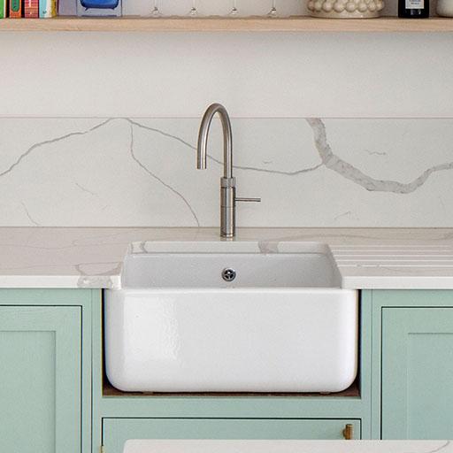 Light blue kitchen cabinets with gold handles, a white countertop, a tall green plant, and wicker bar stools at a marble-topped island in a bright, modern kitchen.