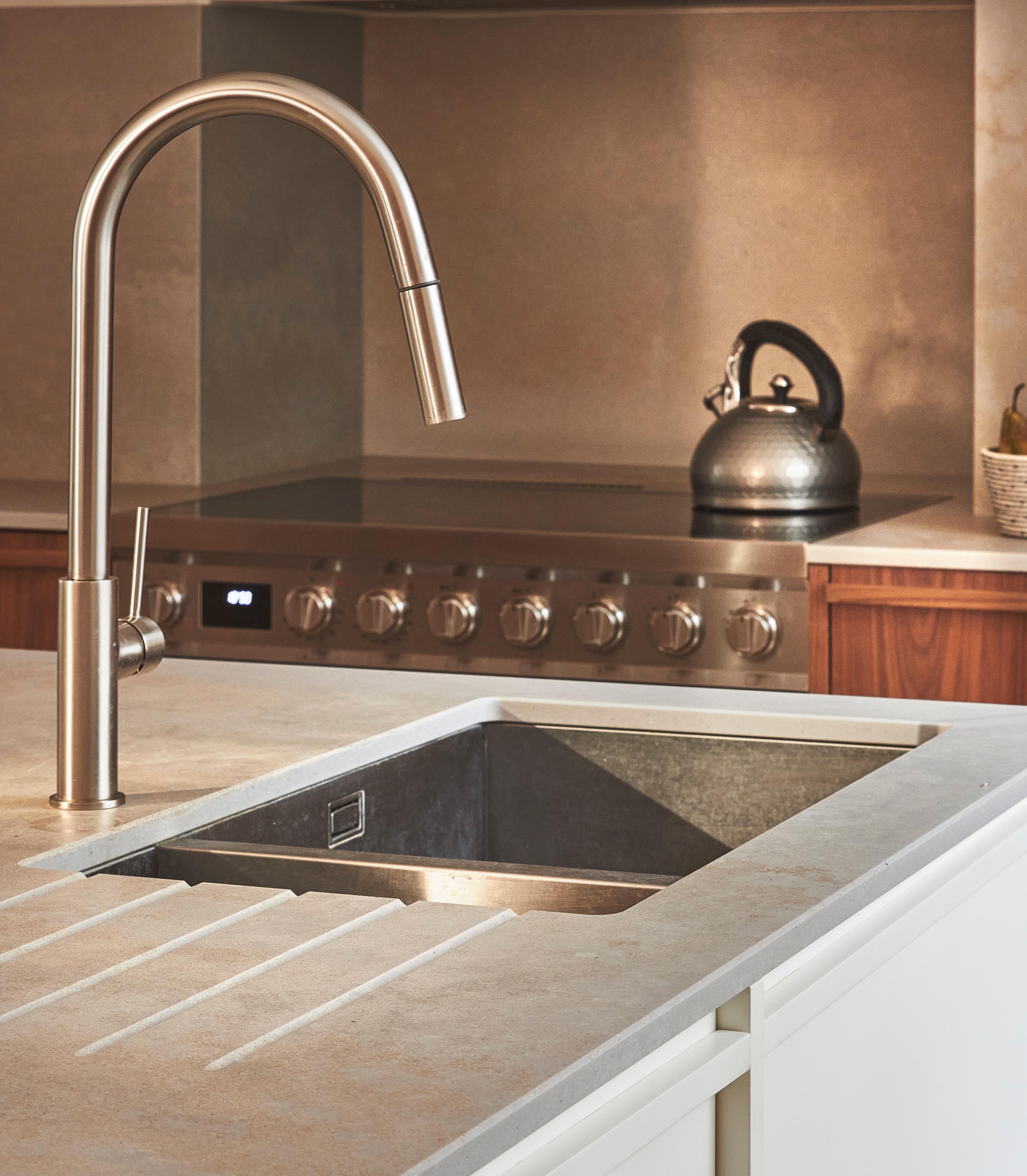 Modern kitchen with a stainless steel sink and sleek faucet in the foreground. An oven with a stovetop and a kettle are visible in the background. The countertops are a light stone, complementing the minimalist decor.