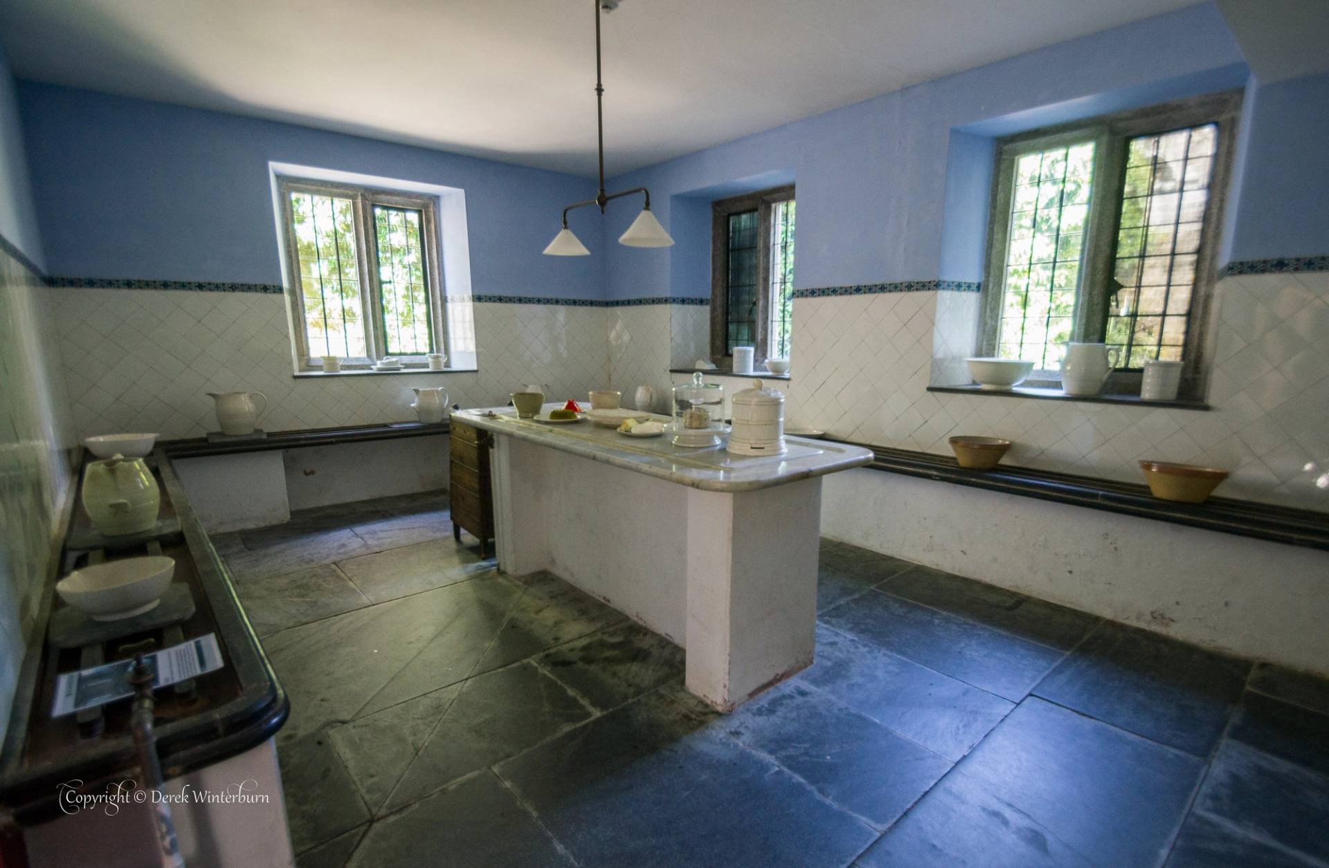 A historic dairy or scullery room featuring blue upper walls, white tiled lower walls, a central stone prep table, and flagstone flooring, with deep-set windows and vintage ceramic bowls and pitchers on display.