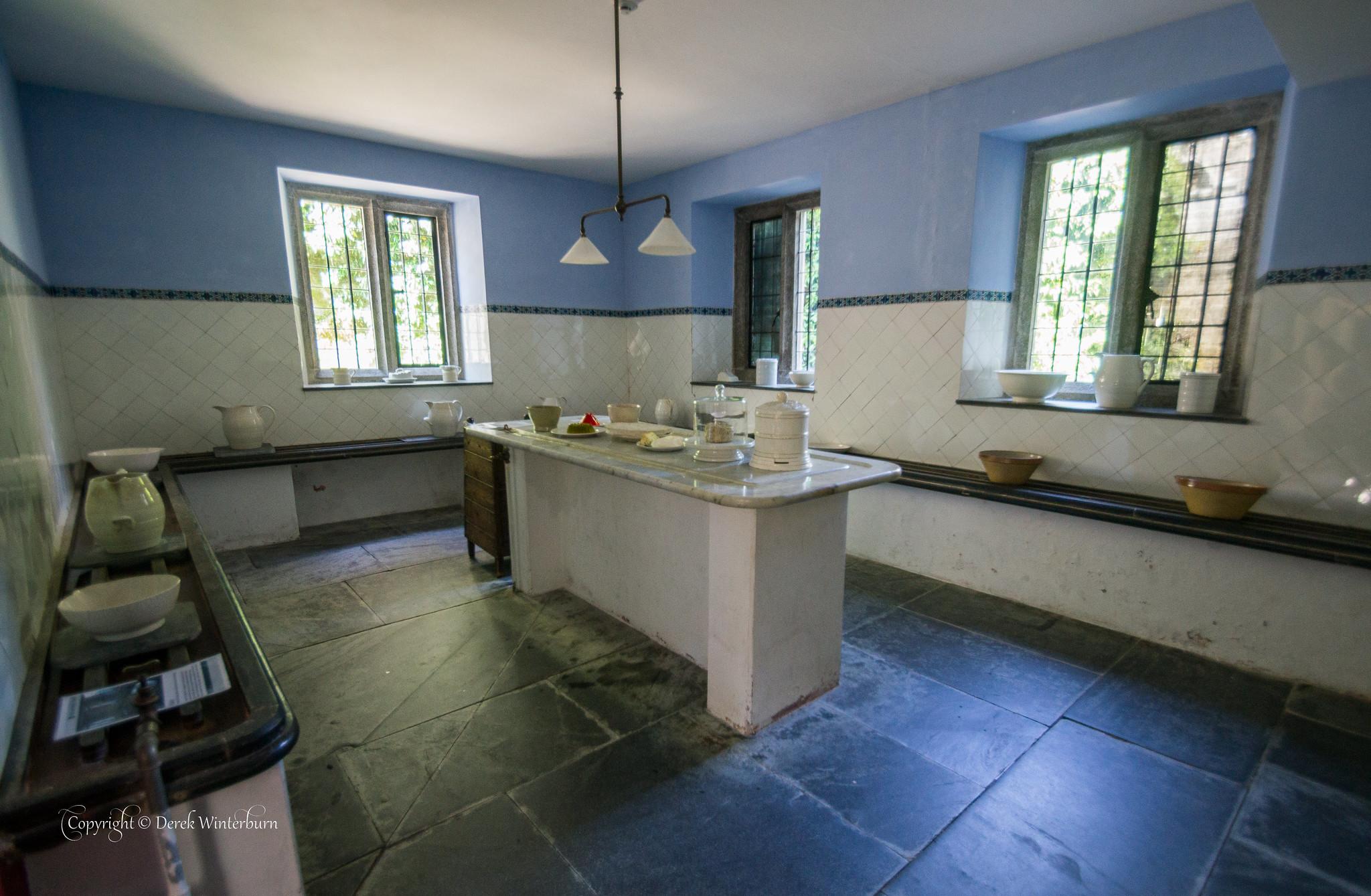 A historic dairy or scullery room featuring blue upper walls, white tiled lower walls, a central stone prep table, and flagstone flooring, with deep-set windows and vintage ceramic bowls and pitchers on display.