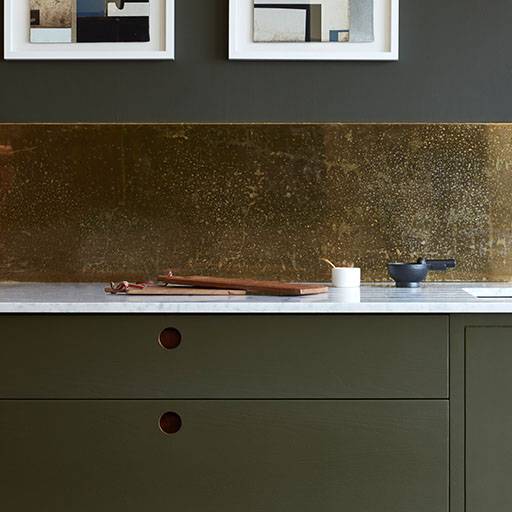 Modern kitchen with matte green cabinets, a white marble countertop, a gold metallic backsplash, a cutting board, a small container, and a dark bowl. Minimalist framed art hangs above the backsplash.