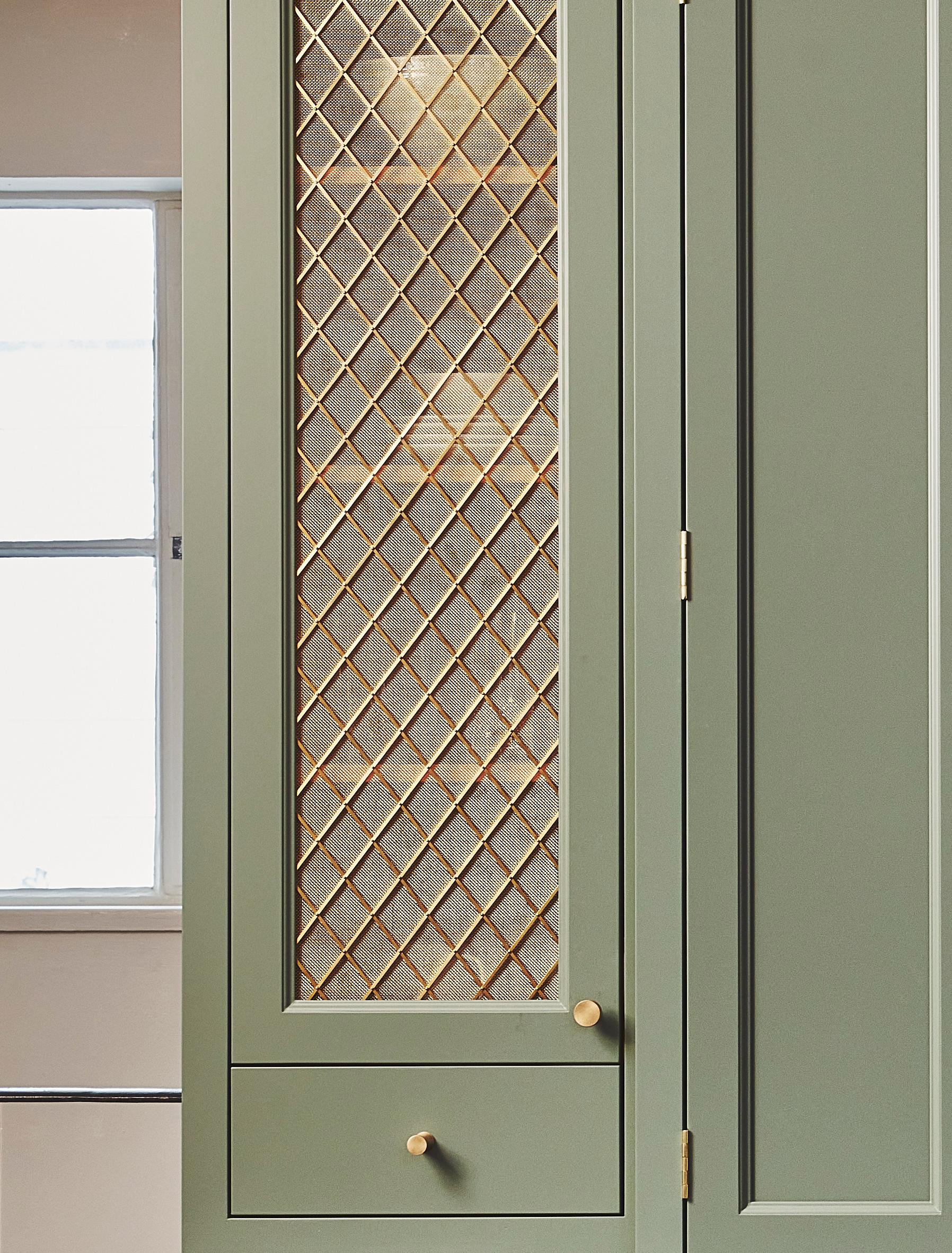 A green cabinet door with a gold mesh lattice panel and two round gold knobs, next to a window with frosted glass letting in natural light.