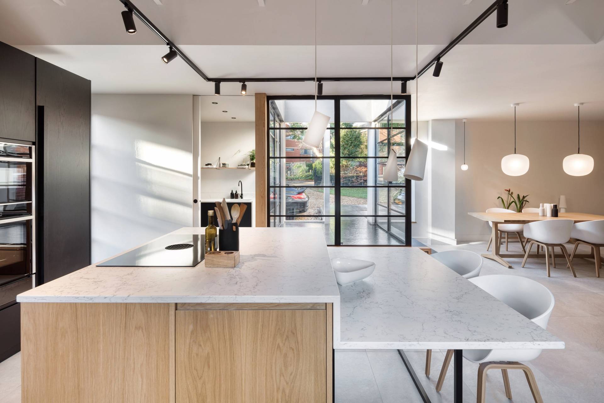 Modern kitchen and dining area with a large island featuring a white marble countertop and light wood paneling. Hanging pendant lights illuminate the space. In the background, a dining table with white chairs sits near large windows.