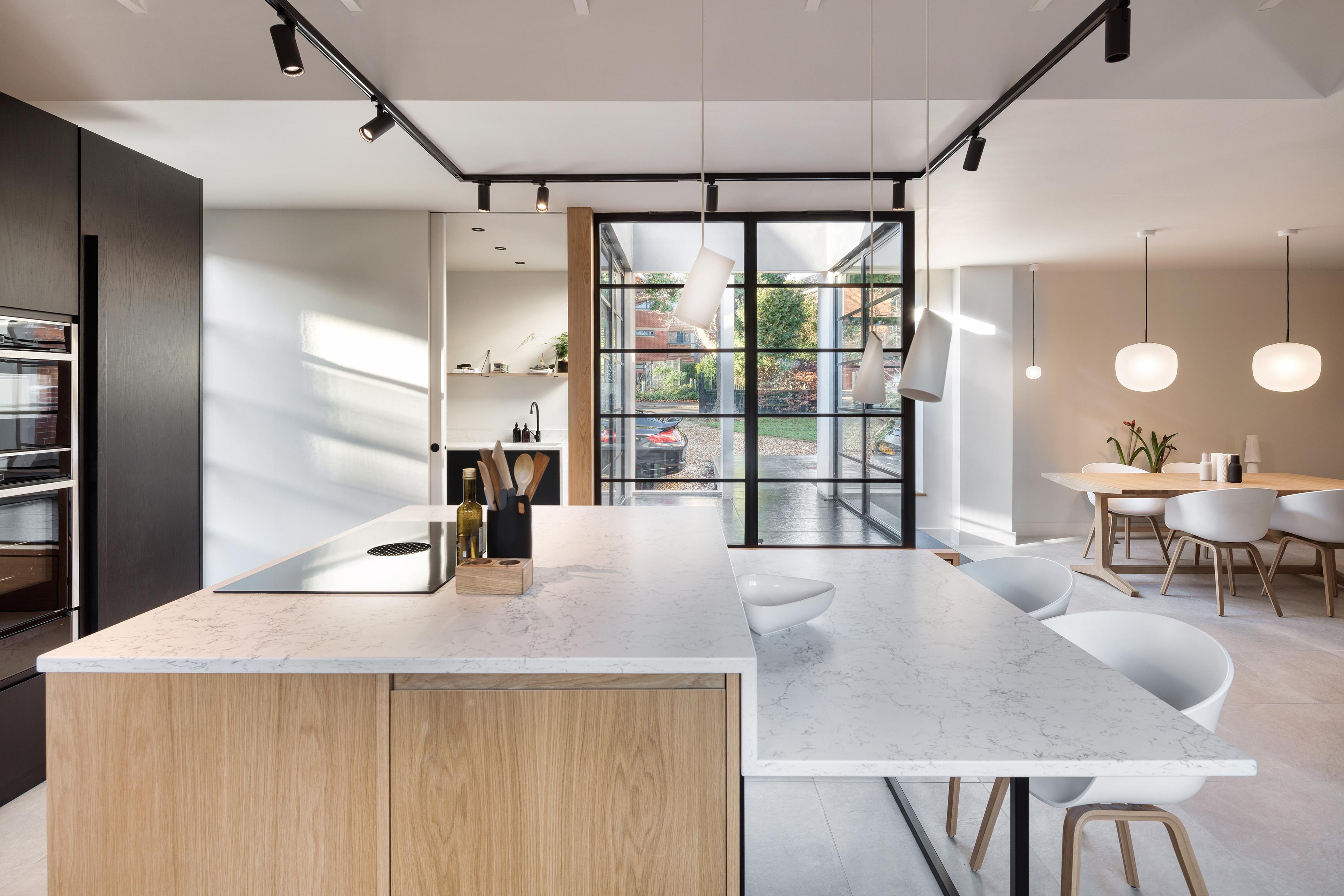 Modern kitchen and dining area with a large island featuring a white marble countertop and light wood paneling. Hanging pendant lights illuminate the space. In the background, a dining table with white chairs sits near large windows.