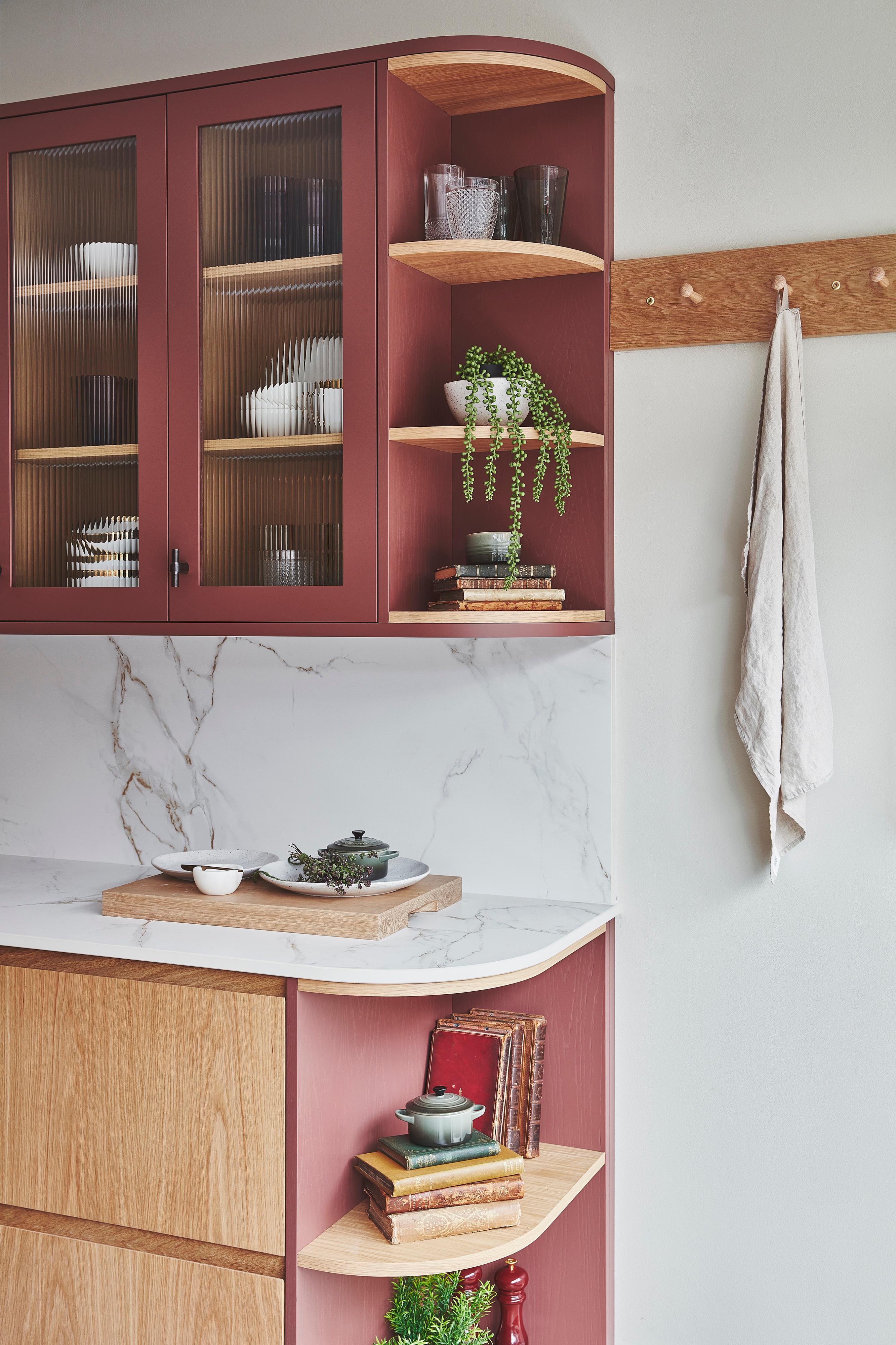 A kitchen corner with wooden cabinets and red shelves. A marble countertop holds a cutting board, bowl, and herbs. Books and potted plants are on the shelves. A wooden coat rack with a white cloth is on the right wall.