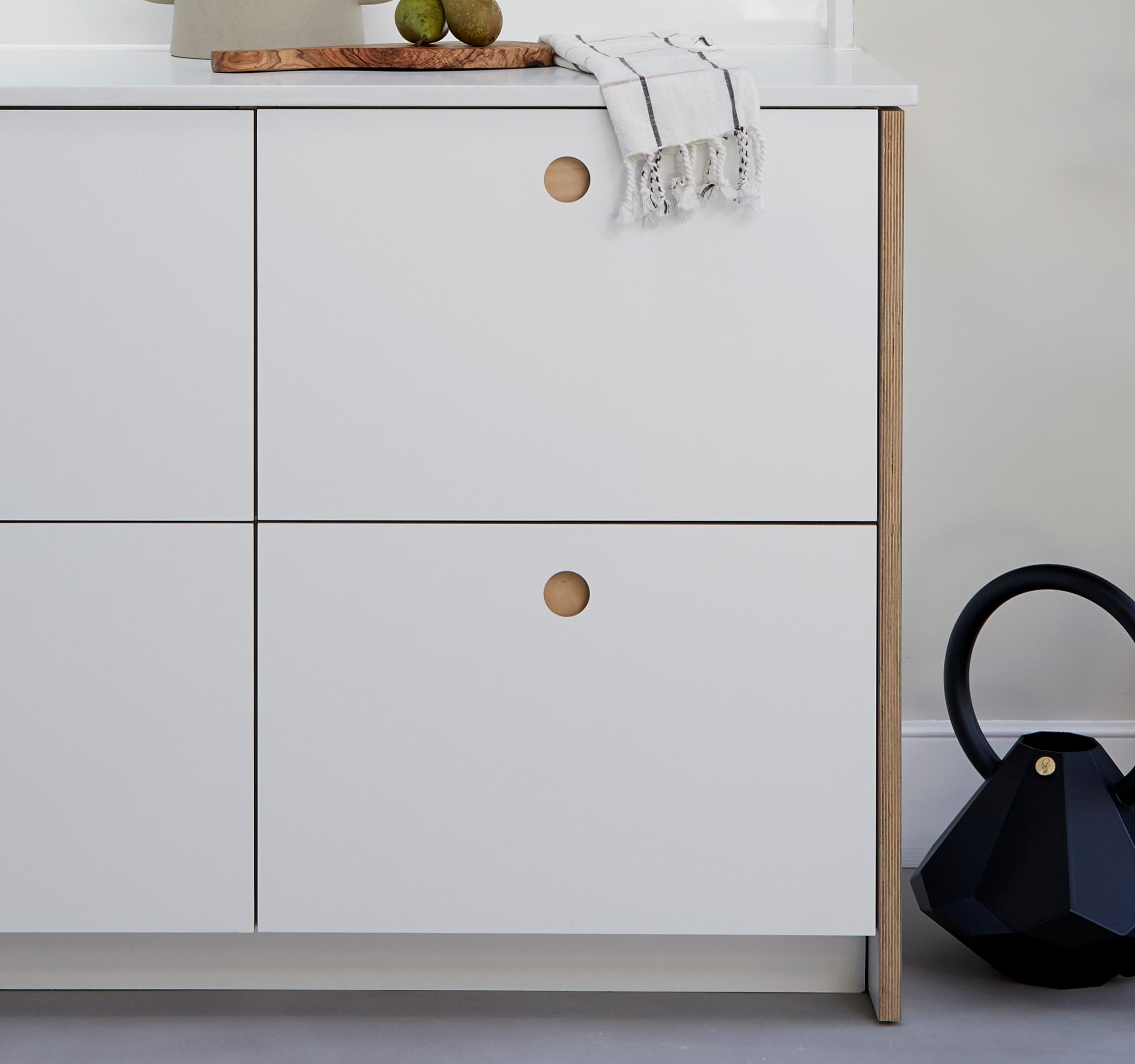A modern white cabinet with four square doors, each featuring a round wooden handle, stands next to a black geometric watering can. A white towel and some fruit rest on top of the cabinet.