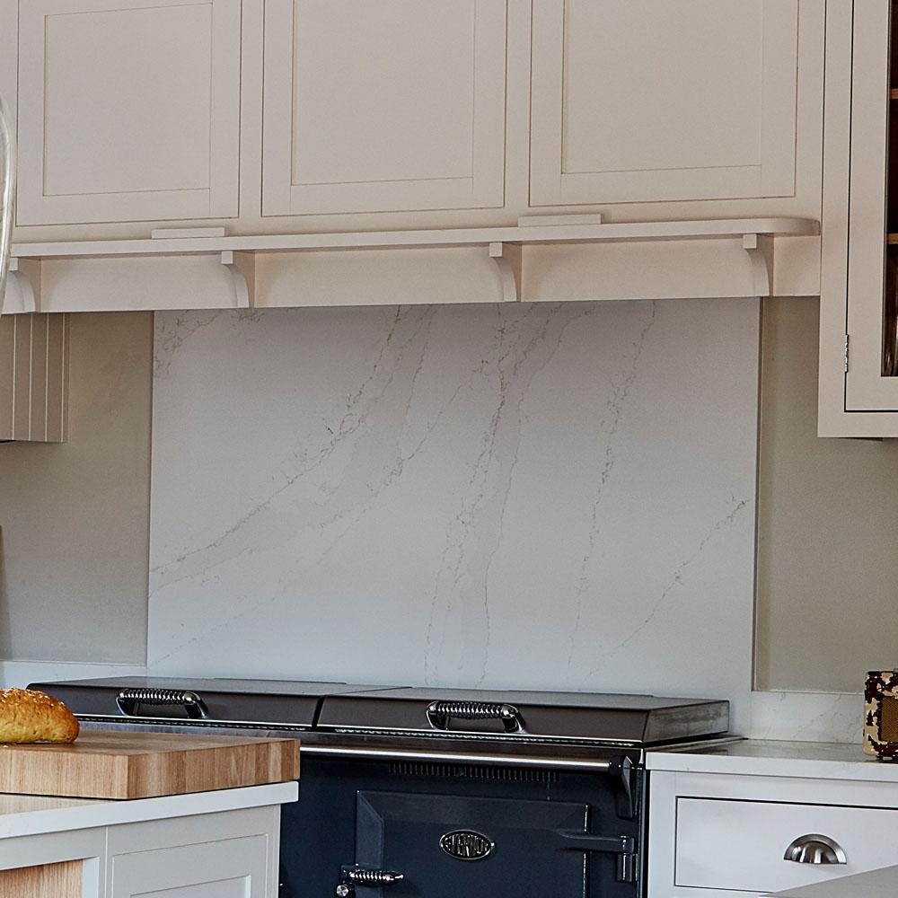 A modern kitchen with light-colored cabinets, a marble backsplash, and a dark-colored stove. A loaf of bread sits on a wooden cutting board on the countertop in the foreground.
