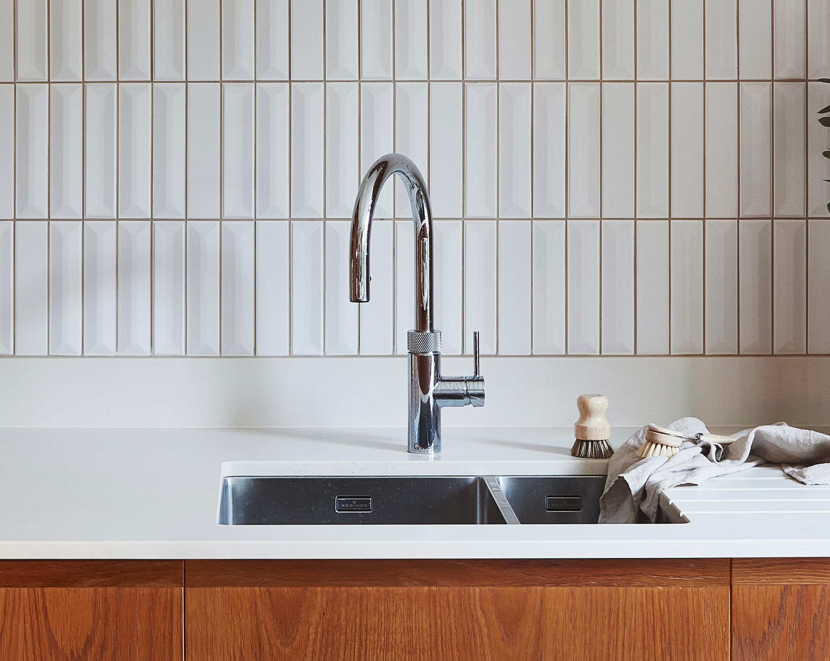 A modern kitchen sink setup with a sleek chrome faucet over a double stainless steel basin. The countertop is white, contrasting with the wooden cabinetry beneath. A dish brush and cloth are placed on the counter. Vertical white tiles are on the backsplash.