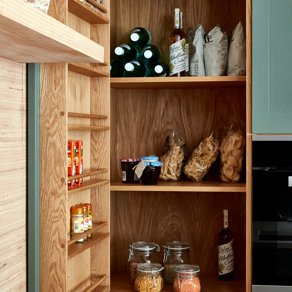 Wooden pantry shelves stocked with bottles, jars, bags of pasta, snacks, and canned goods. Various food items are organized neatly on the shelves, with jars and boxes visible on the left side.