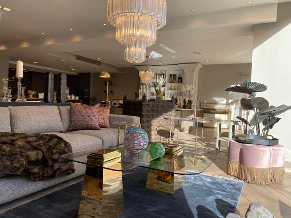 Elegant, sunlit luxury showroom featuring a modern grey sofa, a gold and glass coffee table, a tiered crystal chandelier, and high-end home decor accessories.