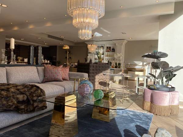 Elegant, sunlit luxury showroom featuring a modern grey sofa, a gold and glass coffee table, a tiered crystal chandelier, and high-end home decor accessories.