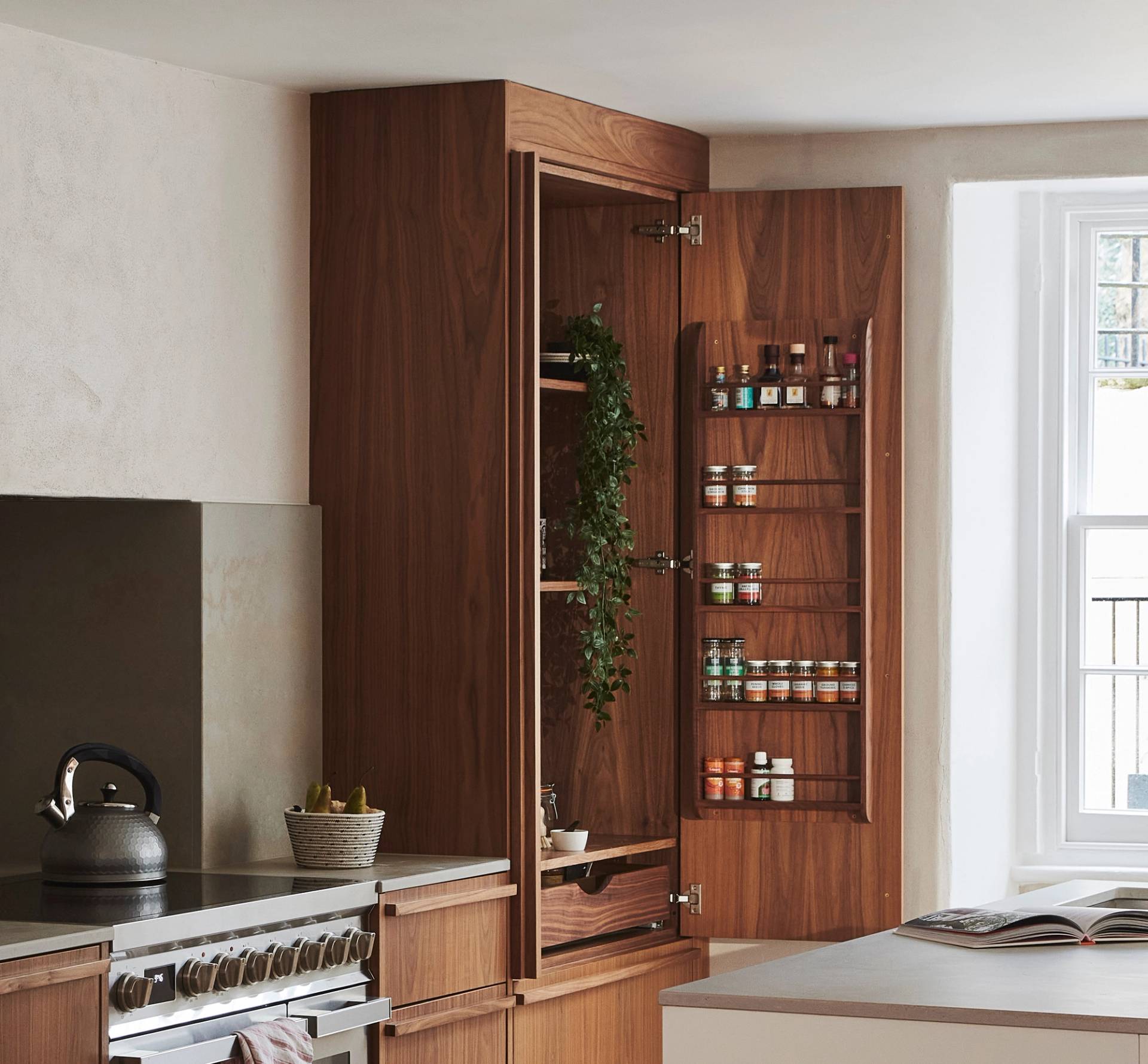 A kitchen with wooden cabinets featuring an open cabinet door. Inside, jars of spices are organized on shelves, and a plant drapes over one shelf. A kettle sits on the stove, and sunlight filters through a window.