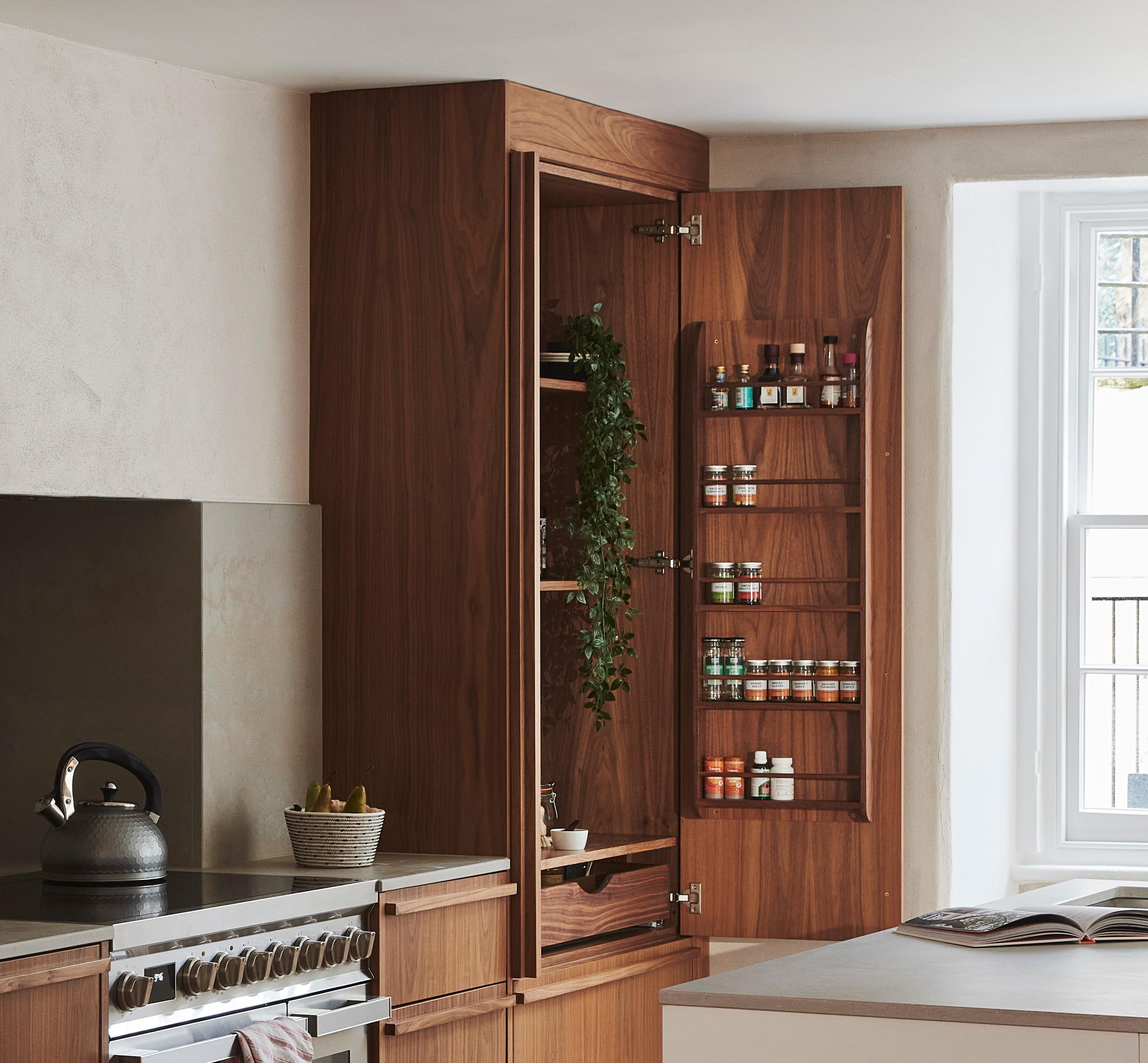 A kitchen with wooden cabinets featuring an open cabinet door. Inside, jars of spices are organized on shelves, and a plant drapes over one shelf. A kettle sits on the stove, and sunlight filters through a window.