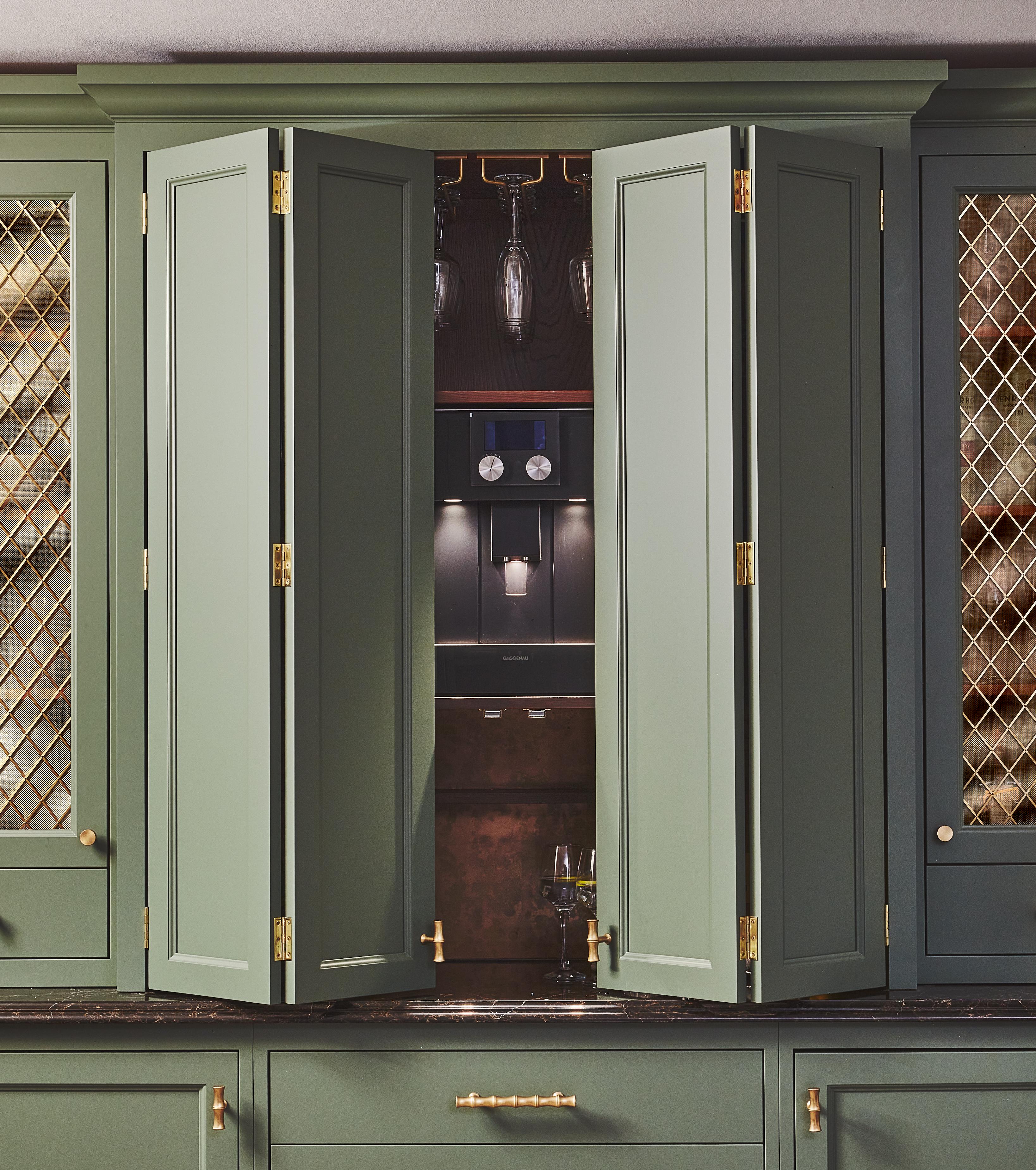 Green kitchen cabinets with gold handles and mesh panels are partially open, revealing a built-in coffee machine, hanging wine glasses, and shelves with glassware inside.