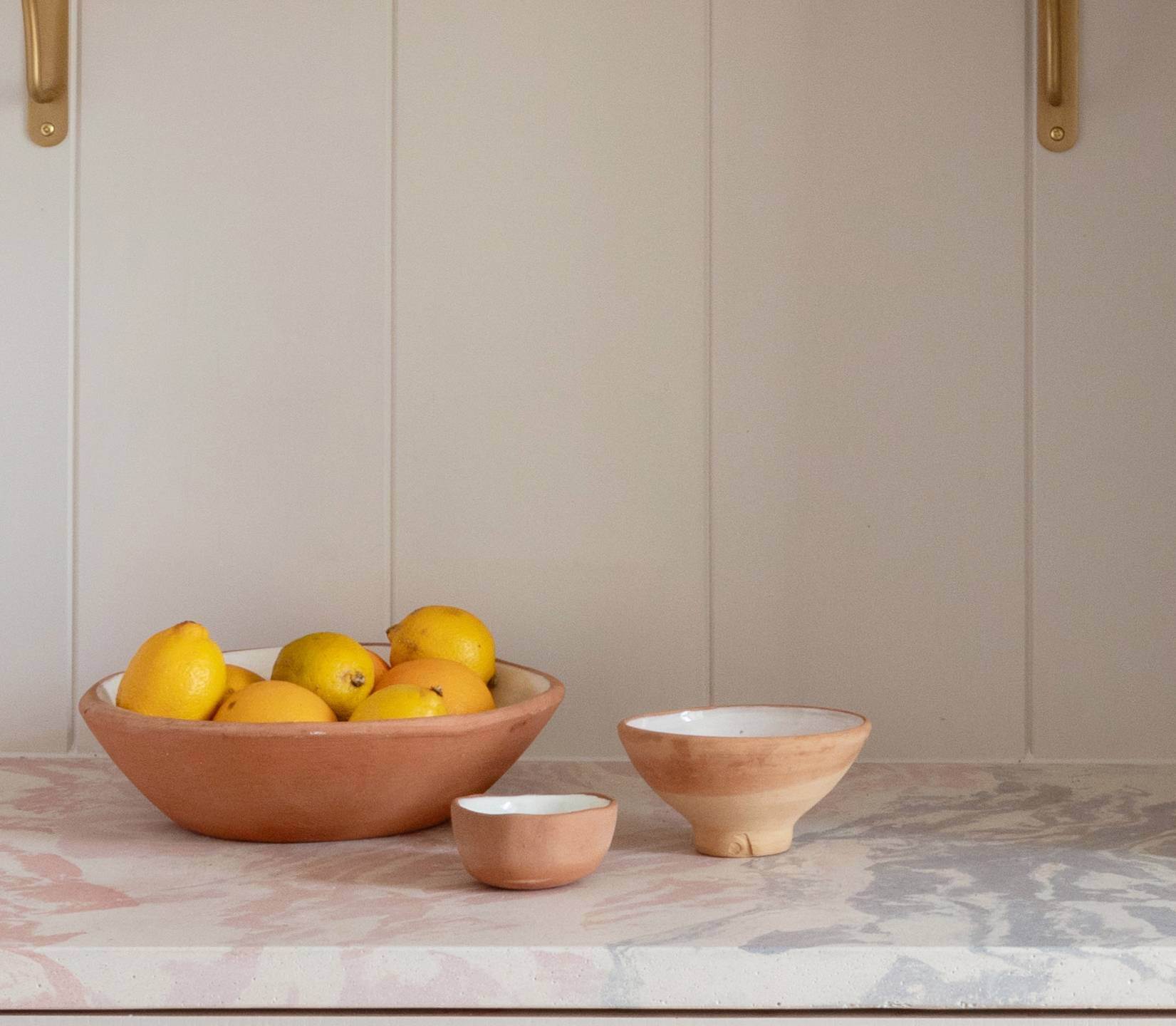 A ceramic bowl filled with yellow lemons sits on a countertop next to two smaller empty ceramic bowls against a white paneled wall.
