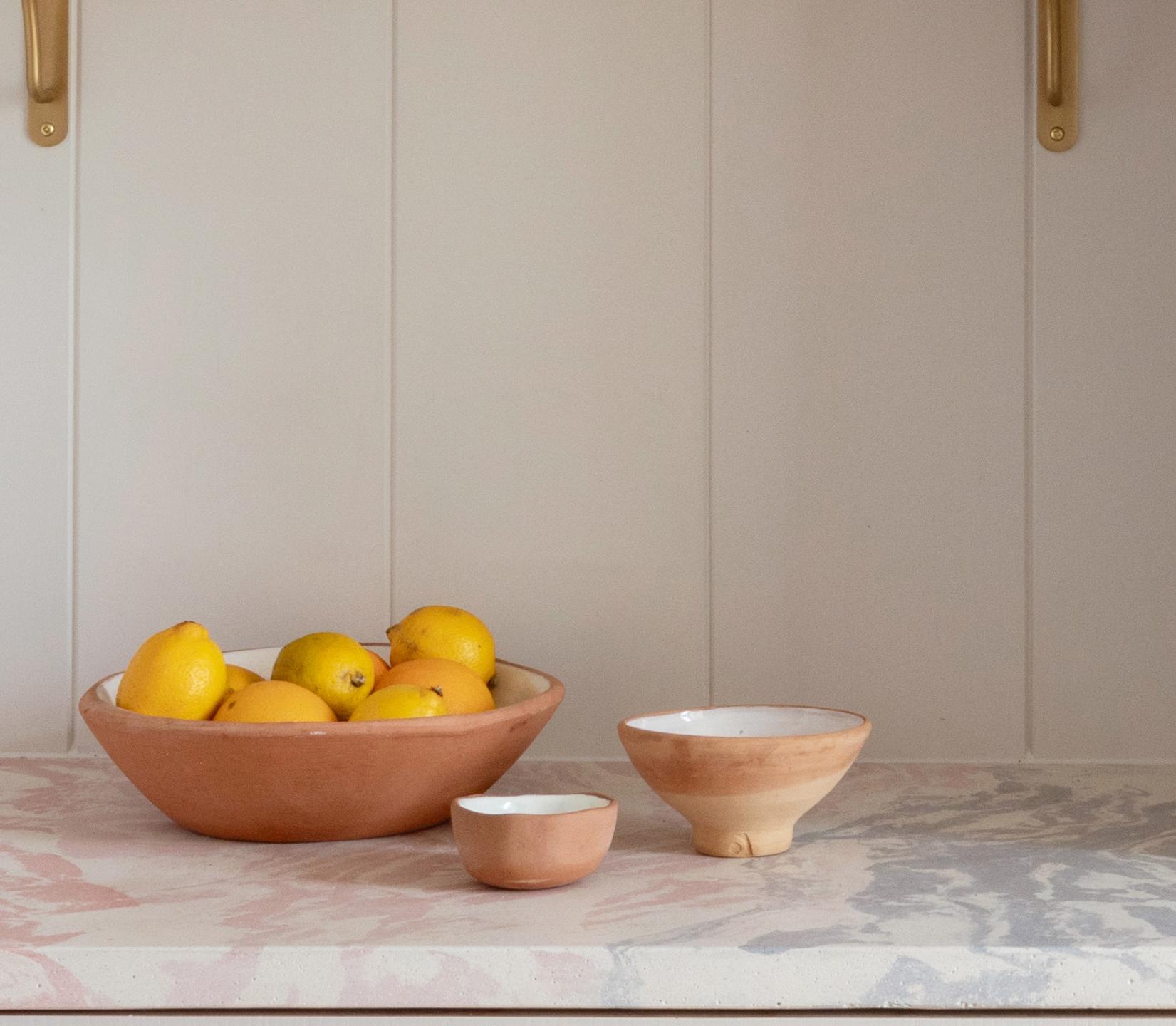 A ceramic bowl filled with yellow lemons sits on a countertop next to two smaller empty ceramic bowls against a white paneled wall.