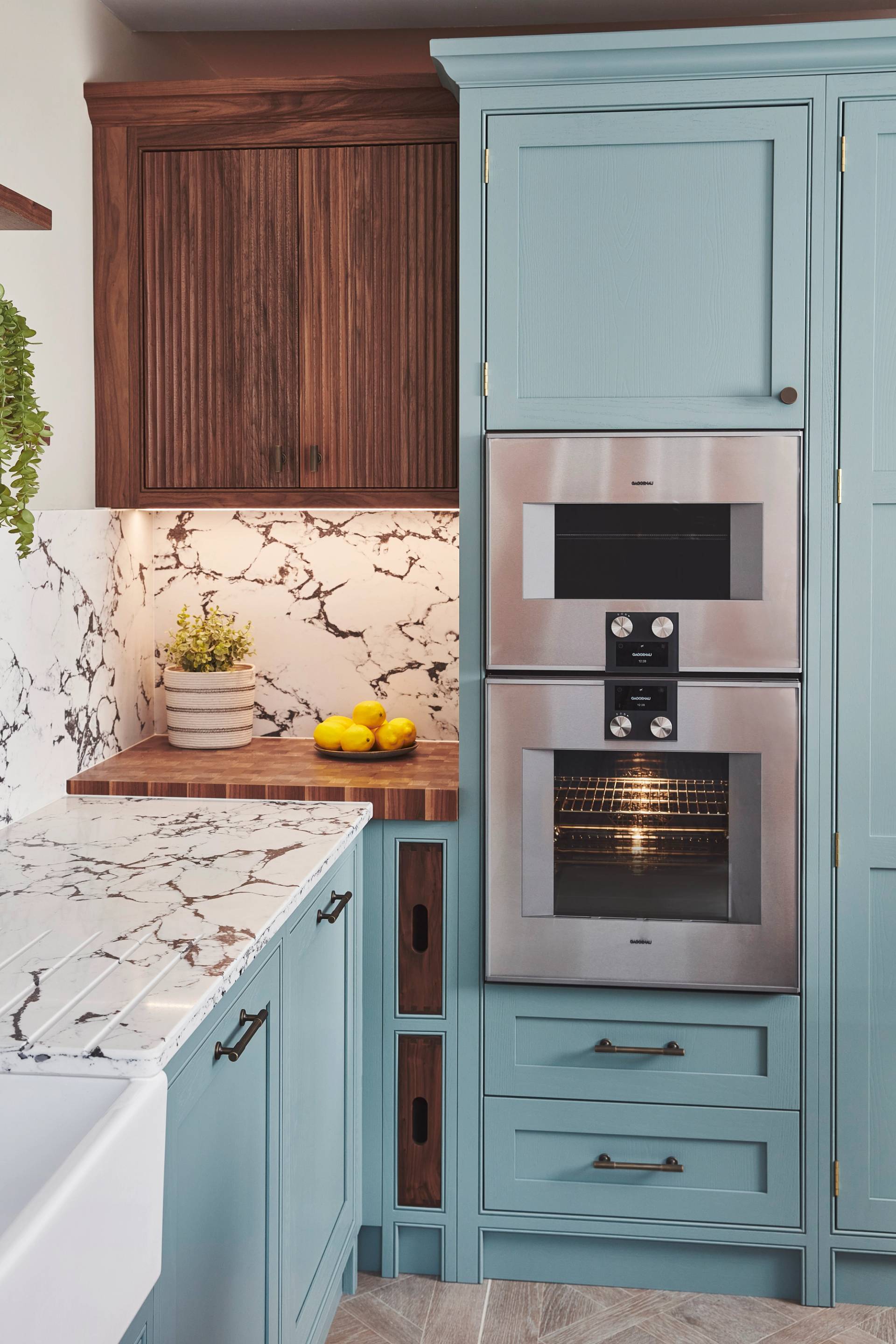 Modern kitchen with a light blue cabinet featuring built-in stainless steel ovens. The countertop and backsplash have a white and gray marble pattern. A small wooden section holds lemons, a plant, and features an underlit dark wooden cabinet.