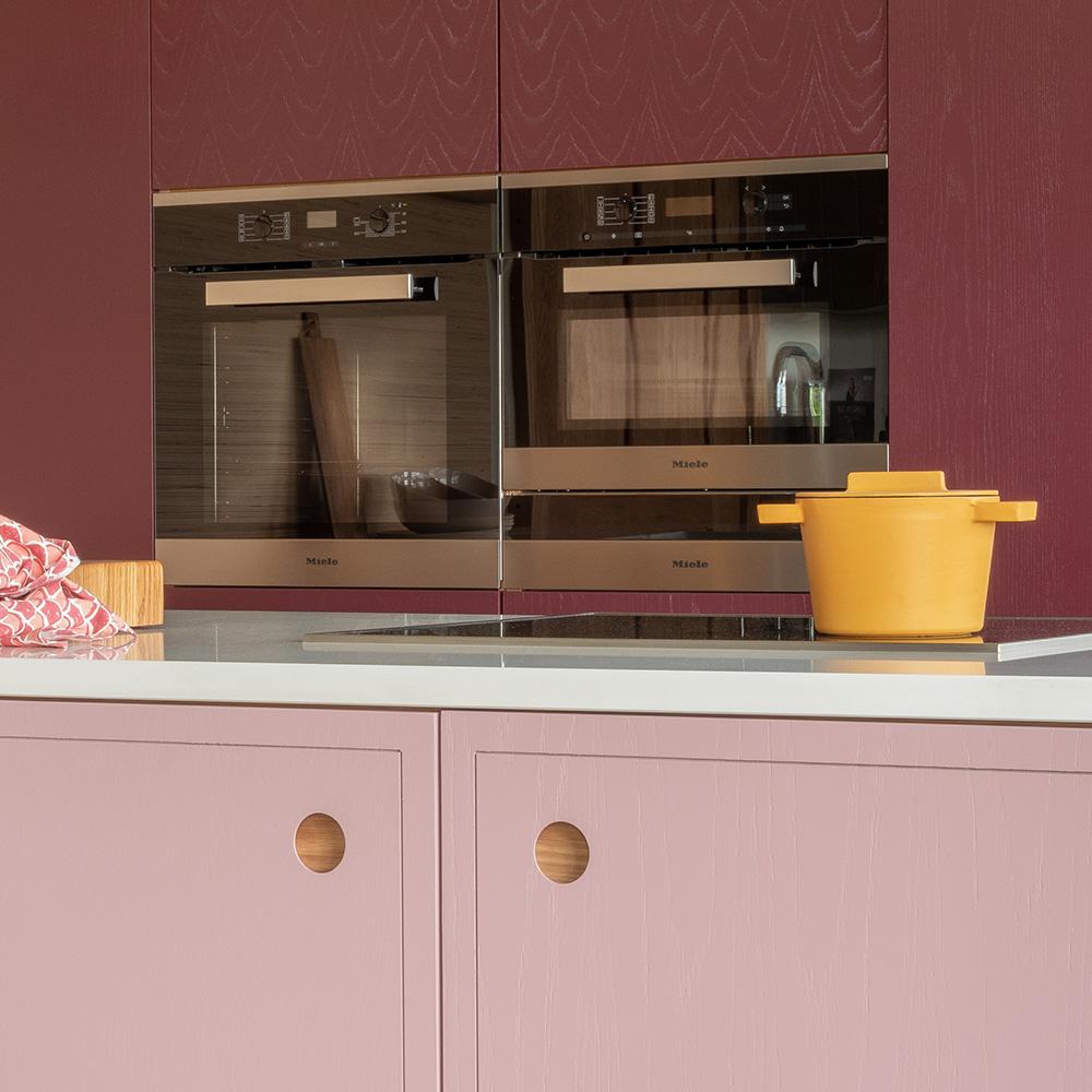 A modern kitchen with maroon cabinets, built-in stainless steel ovens, a pink lower cabinet with round handles, a yellow pot on the white countertop, and a red patterned kitchen towel.